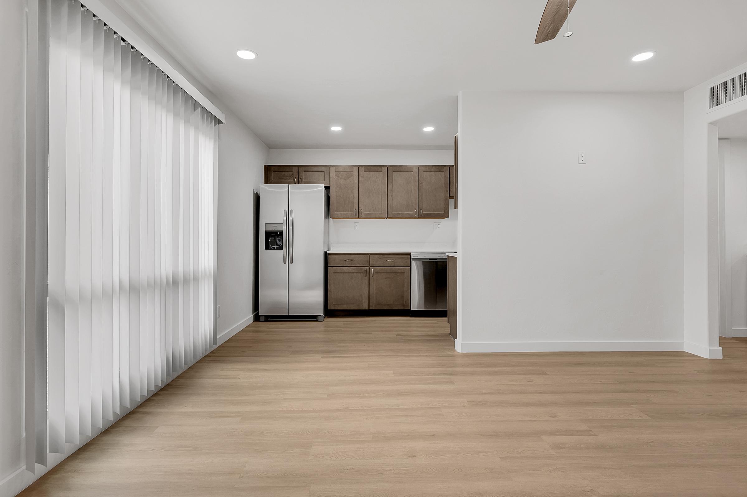 Modern kitchen and living area featuring light wood flooring, a stainless steel refrigerator, and dark wood cabinetry. The space is well-lit with recessed lighting and large vertical blinds on the windows, creating an airy and contemporary atmosphere.