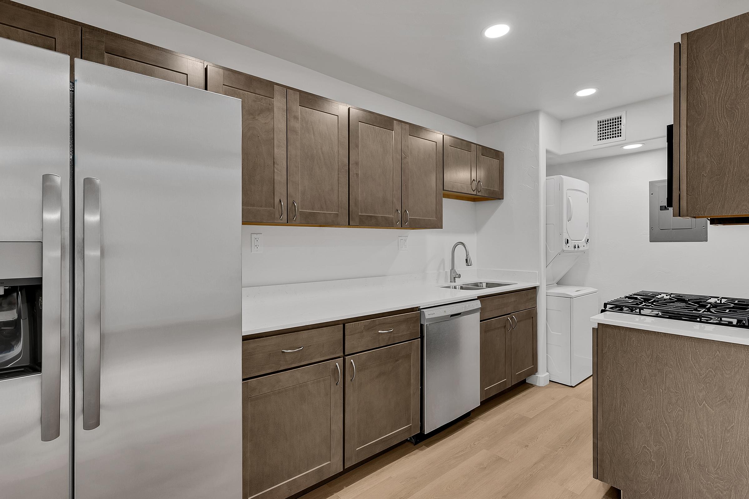 Modern kitchen featuring stainless steel appliances, including a refrigerator and dishwasher. It has wooden cabinets, white countertops, and a double sink. The space is well-lit with recessed lighting. In the background, there's a stacked washer and dryer unit, and large cabinets provide additional storage.