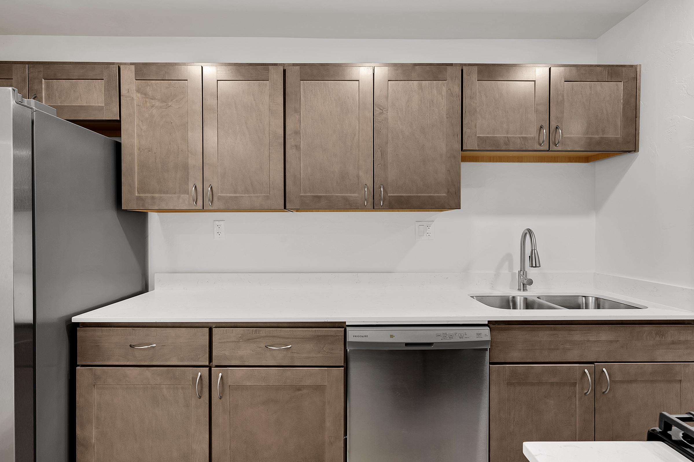 Modern kitchen design featuring wooden cabinets with a light finish, a stainless steel refrigerator, a double sink with a sleek faucet, and a dishwasher. The countertop is white and smooth, providing a clean and contemporary look. The walls are painted white, enhancing the overall brightness of the space.