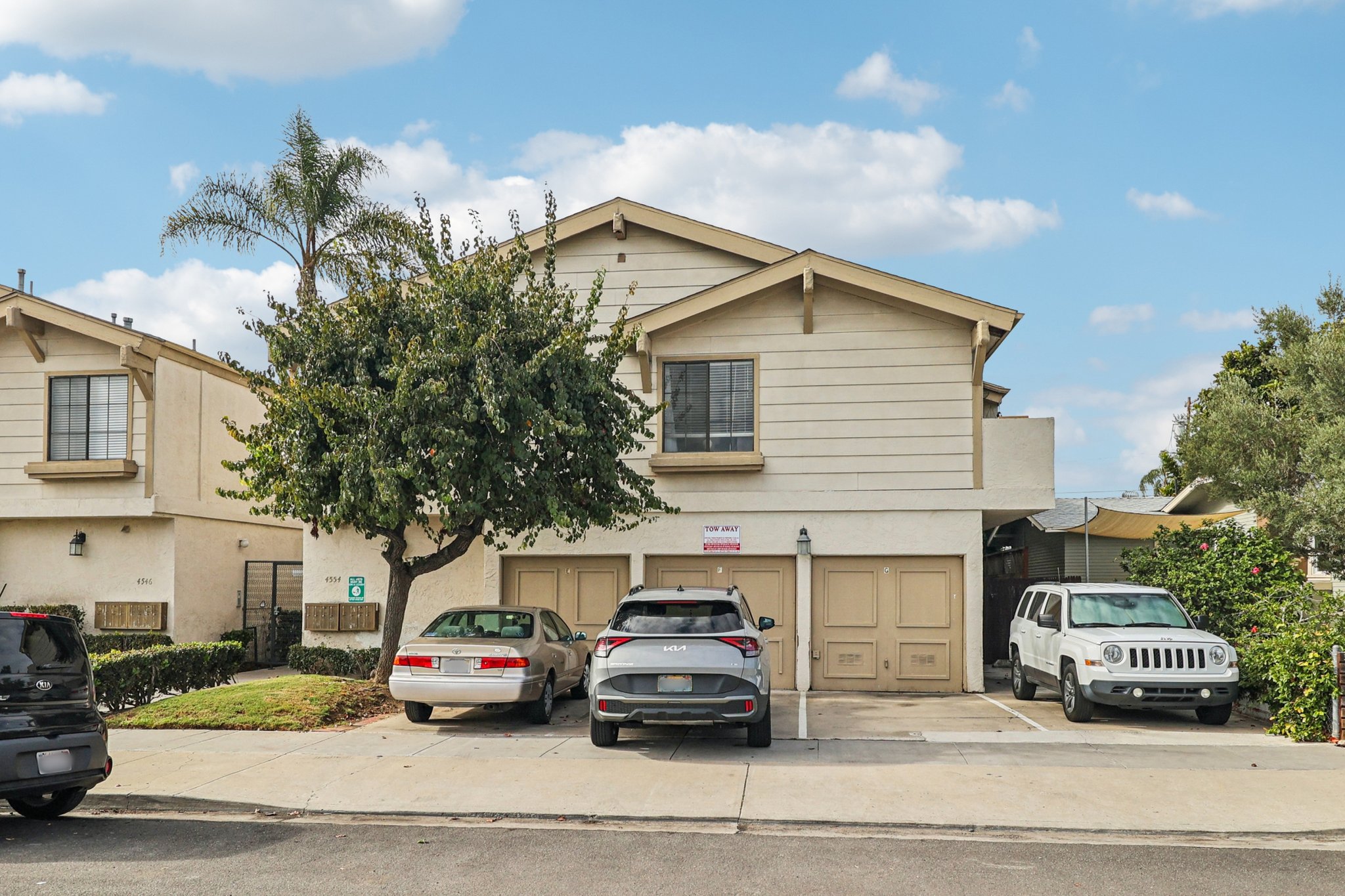 A two-story residential building with beige exterior, featuring a garage and parked vehicles. A palm tree is visible in the foreground, and the sky is partly cloudy. The building has a "For Rent" sign on the front.