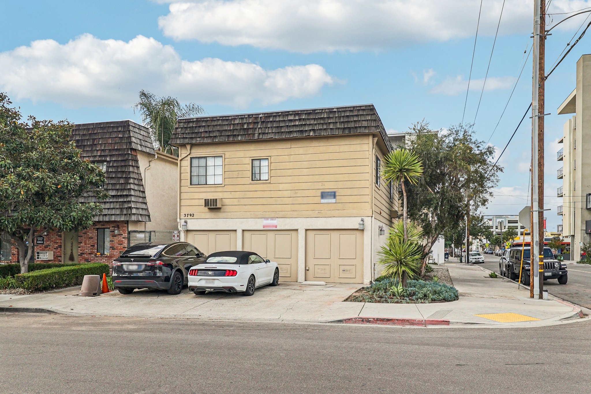 A two-story residential building with a beige exterior and a sloped roof, situated on a corner lot. Two cars are parked in front—one is a black sedan and the other a white sports car. Surrounding greenery includes small trees and shrubs, with a clear blue sky and scattered clouds above.