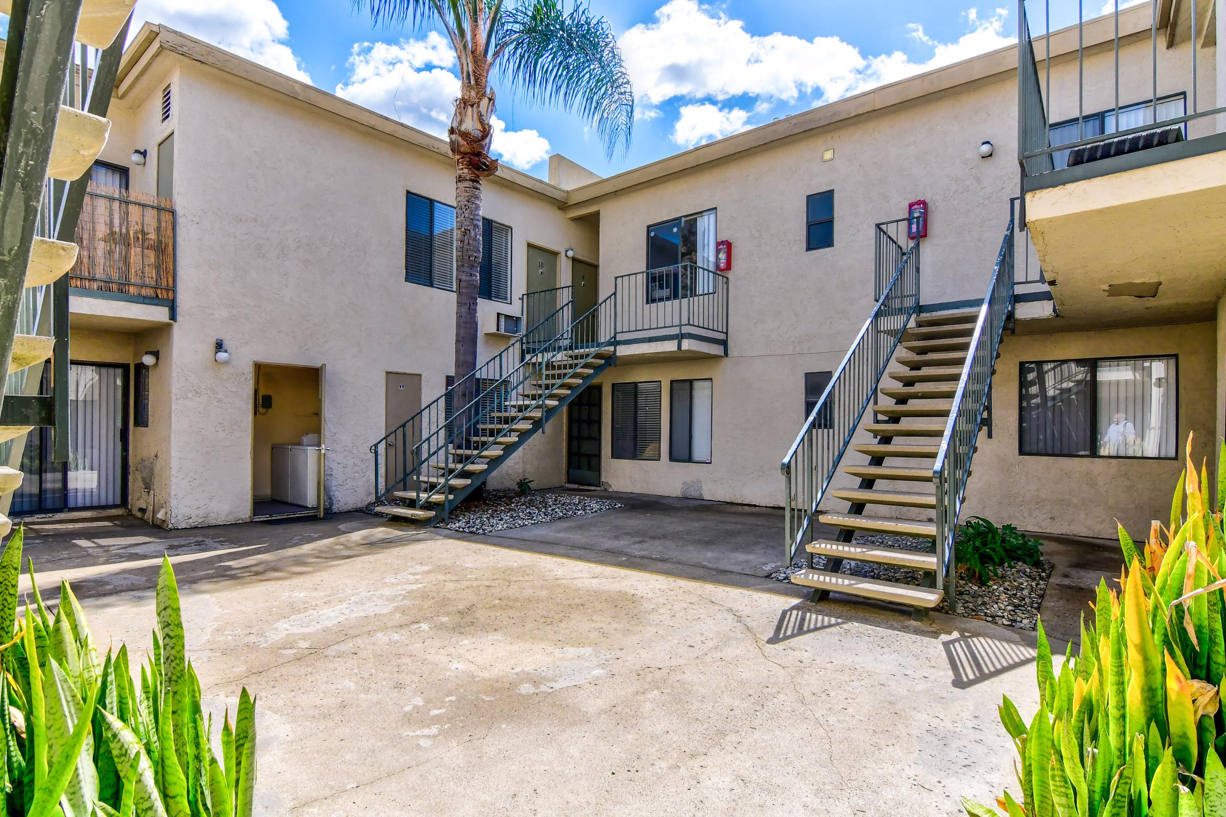 A view of an apartment courtyard with two staircases leading to upper units. The building has palm trees and shrubs in the landscaping, with a paved area between the staircases. Bright blue sky and clouds are visible above.