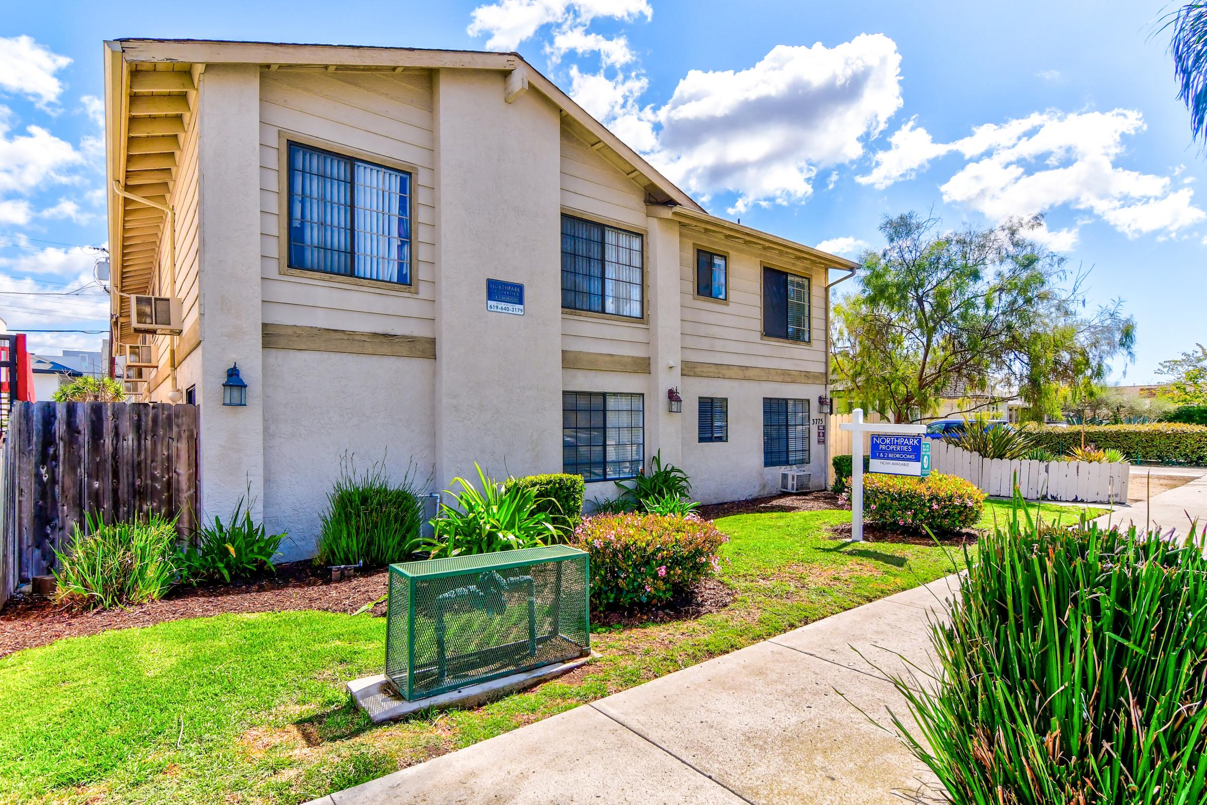 Two-story residential building with a light-colored exterior featuring multiple windows. The front yard has well-maintained grass, colorful shrubs, and a decorative fence. There is a sign in front, indicating the property. The sky is bright with scattered clouds.