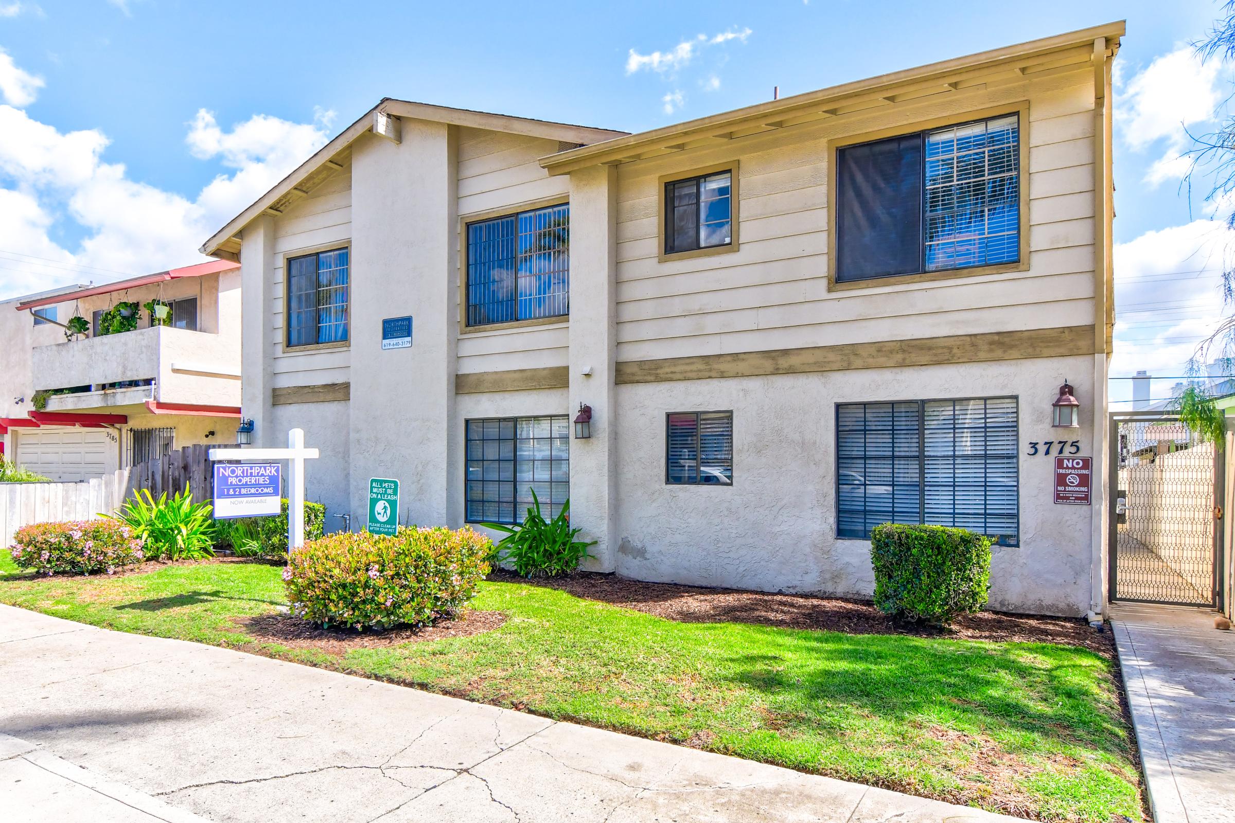 A two-story residential building with a light-colored exterior and multiple windows. The front yard features neatly trimmed bushes and grass. A sign for "Northpoint Apartments" is visible in front, along with a pathway leading to the entrance. The sky is bright and partly cloudy.