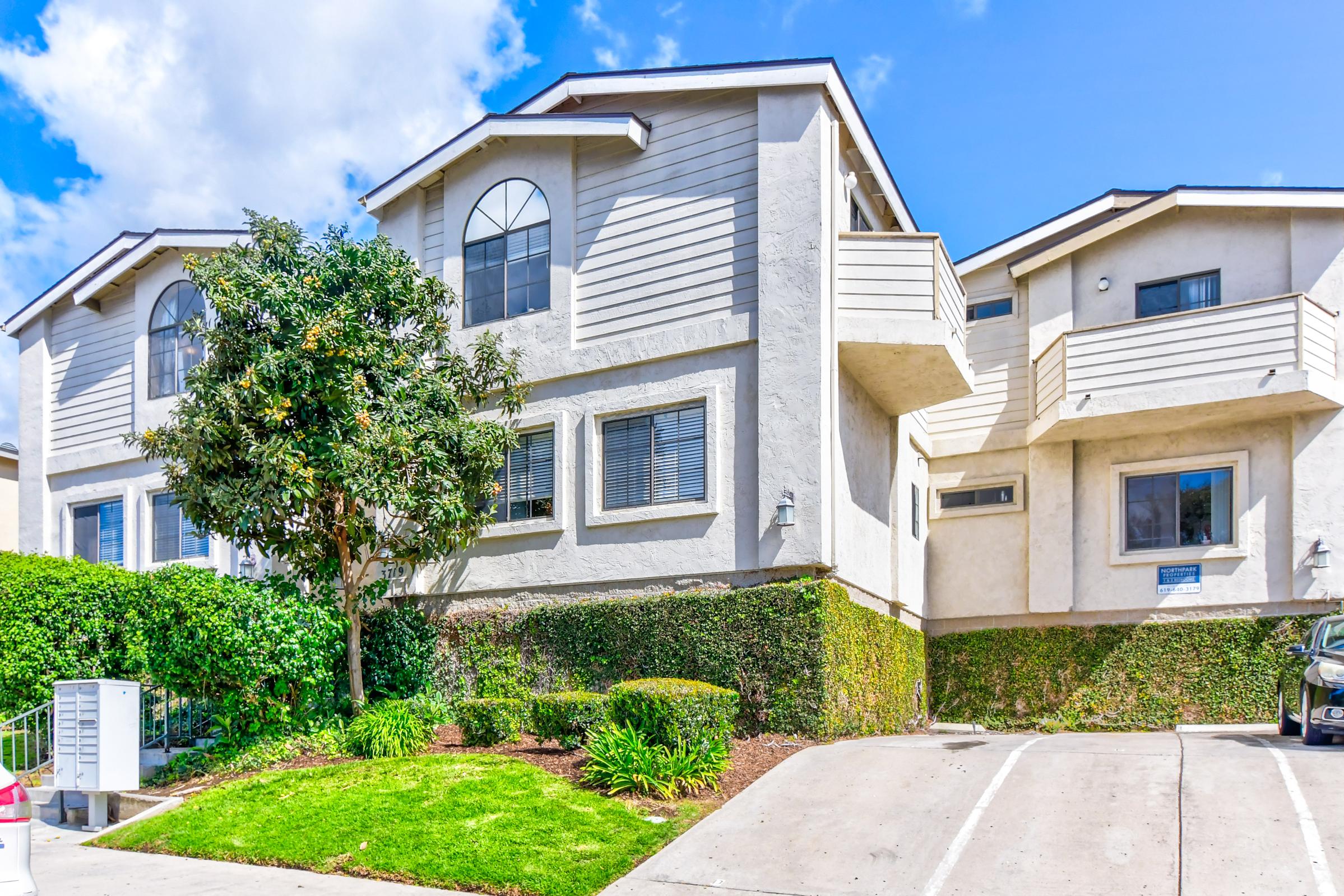 A modern two-story building with a light-colored exterior, featuring large windows and balconies. The surrounding area includes well-maintained greenery, including a small tree and shrubs. A paved parking area is visible in front of the building, with a clear blue sky overhead.