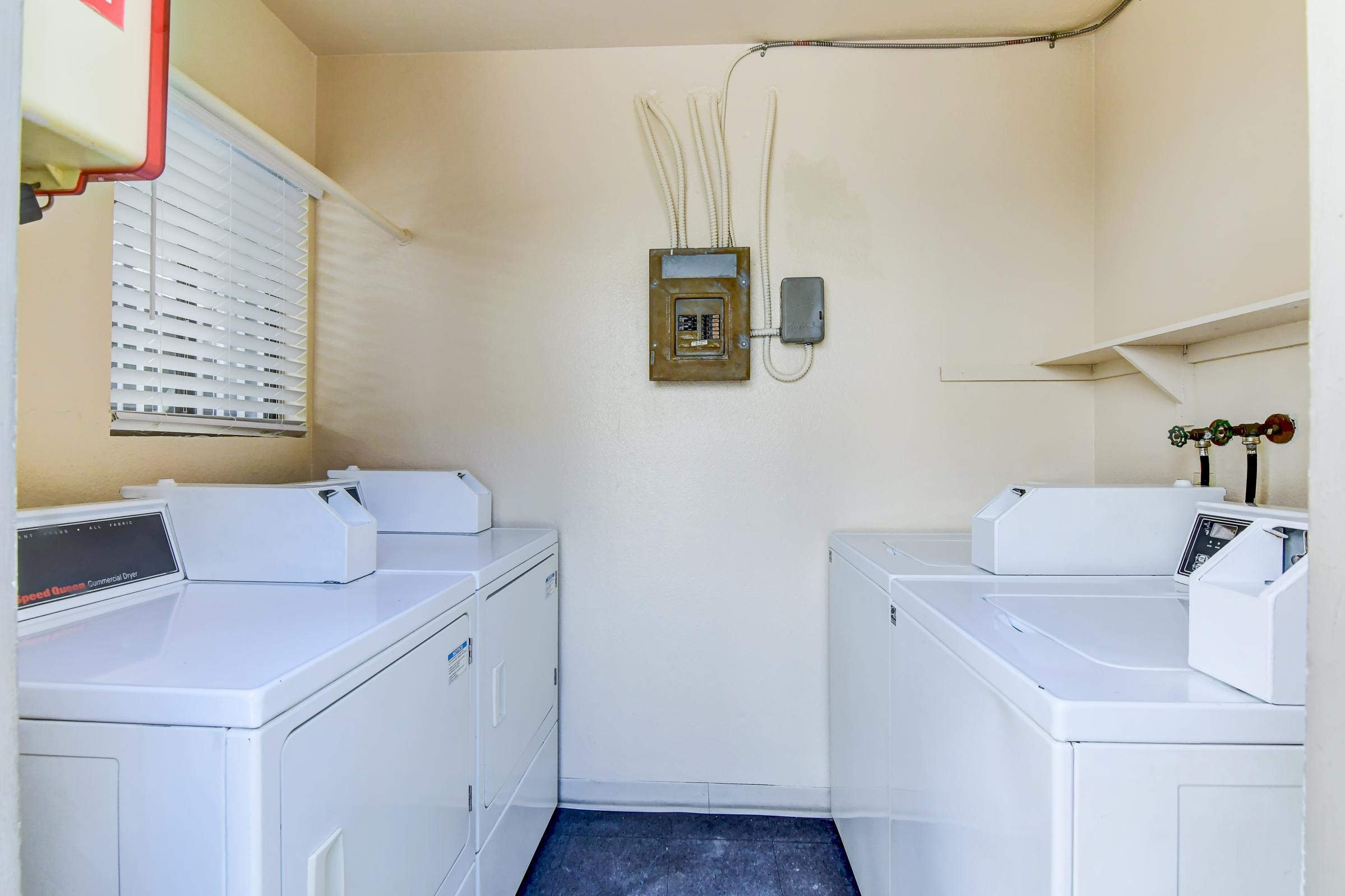 A clean laundry room featuring two white washing machines and a white dryer arranged in a bright, light-colored space. A window with blinds lets in natural light, and a utility panel is mounted on the wall. A shelf is visible above the machines, providing additional storage space.