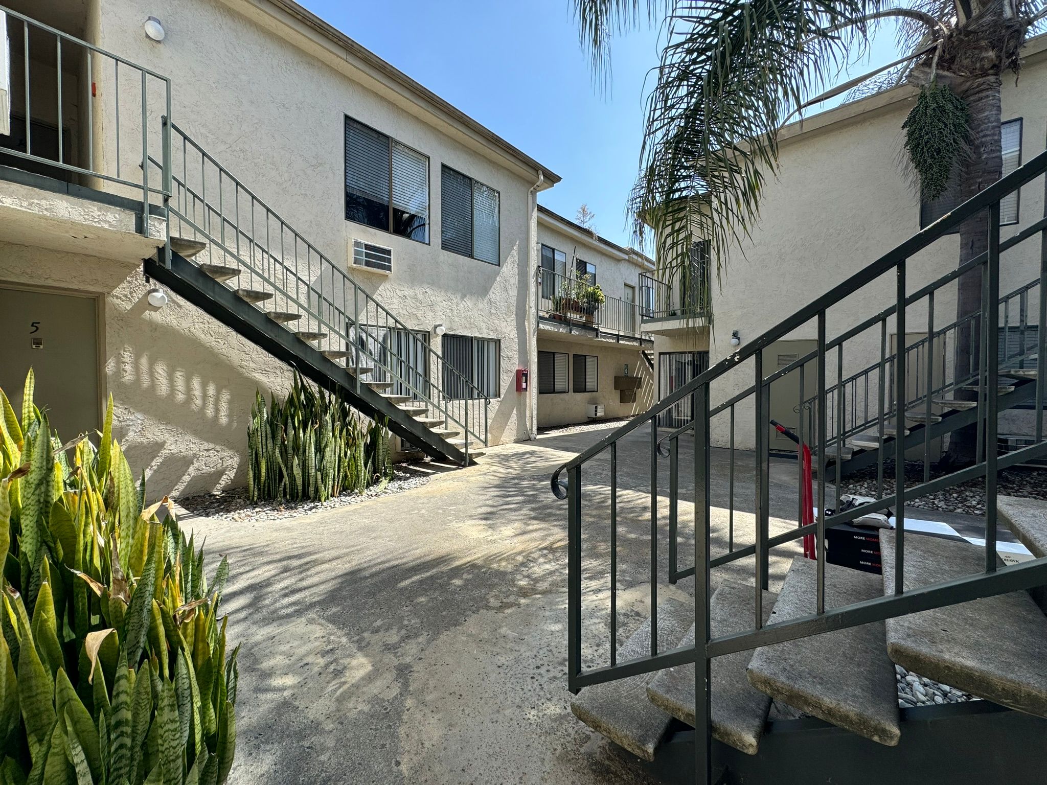 A view of a courtyard in an apartment complex featuring two-story buildings. There are stairs leading to different entrances, palm trees, and decorative plants along the pathway. The sky is clear and blue, creating a bright, welcoming atmosphere.
