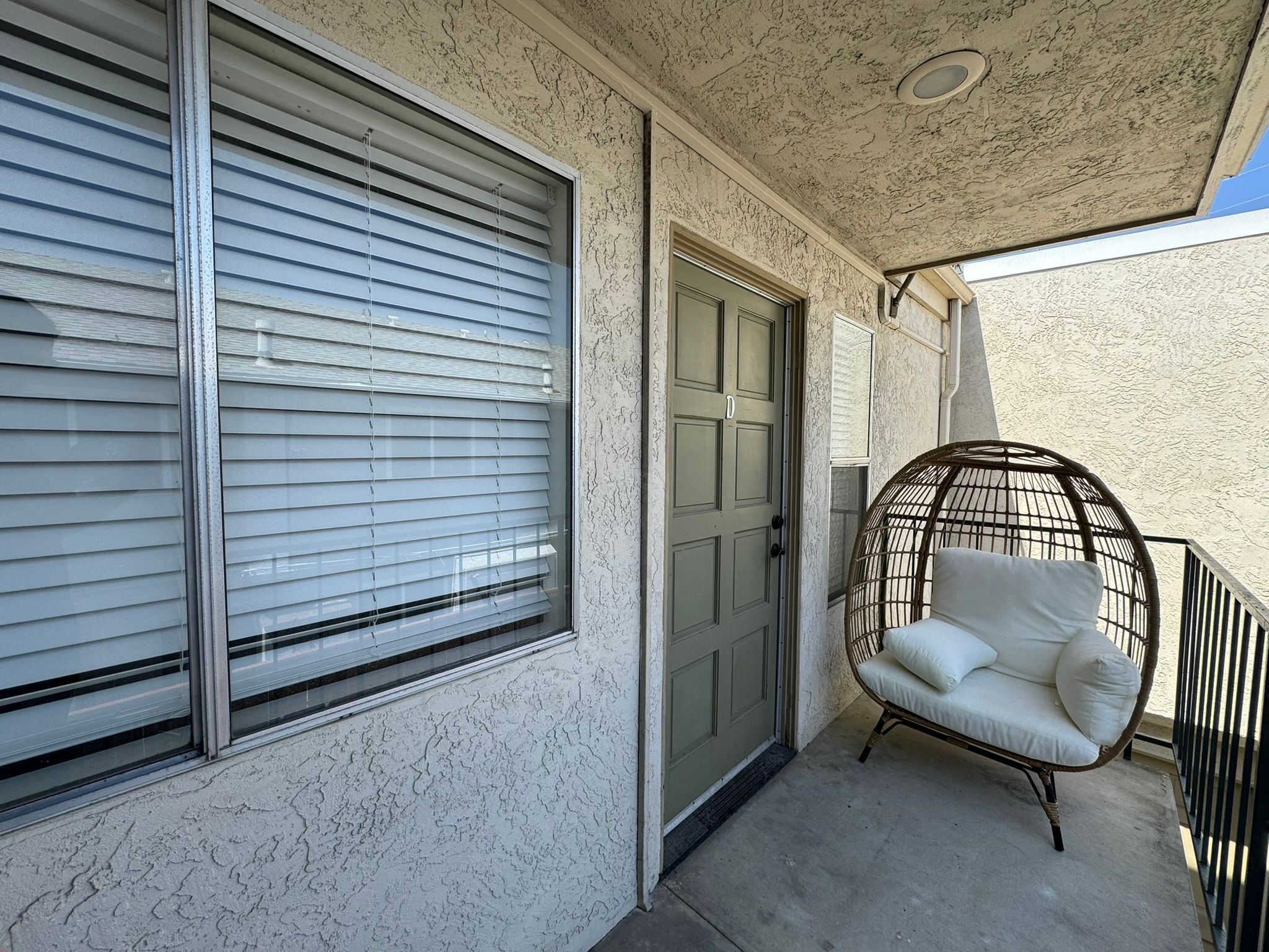 A small outdoor porch area featuring a light gray door, a window with white blinds, and a round, woven chair with a white cushion. The wall has a textured finish, and the area is well-lit by natural sunlight.