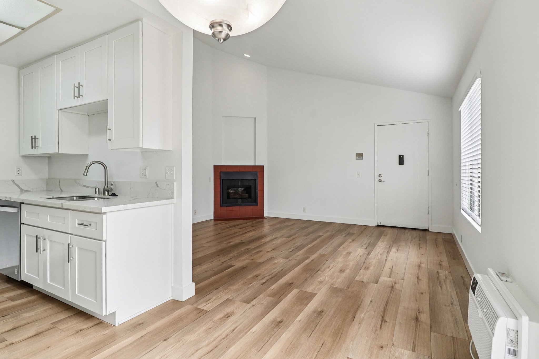 A modern, open-concept living space featuring a kitchen area with white cabinets and a sink, adjacent to a cozy living room with a fireplace. The room has light wood flooring, a large window with blinds, and a front door visible in the background. Walls are painted in a neutral tone, creating a bright atmosphere.