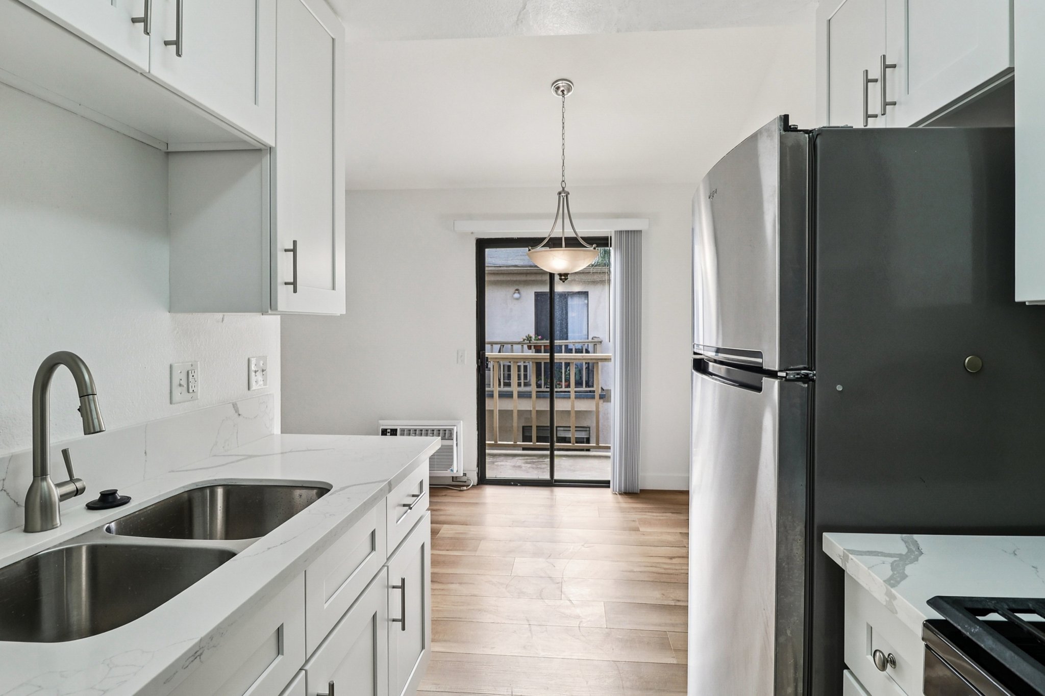 Modern kitchen featuring white cabinetry, a double sink, and stainless steel appliances. Natural light streams in through a sliding glass door leading to a porch. Stylish pendant light hangs above the counter, and hardwood floors add warmth to the space.