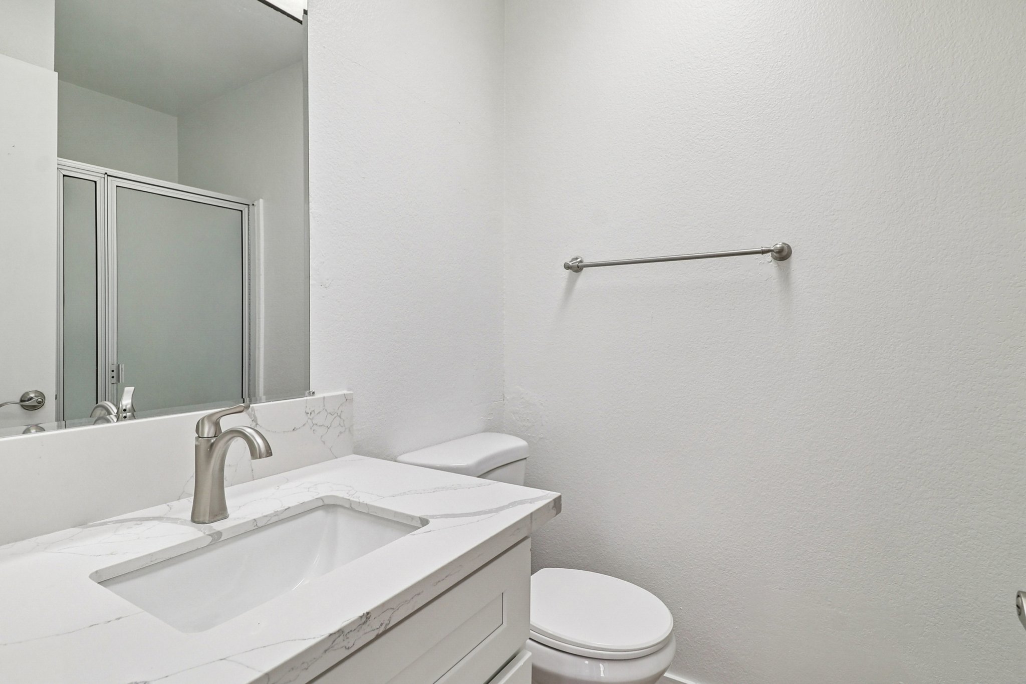 A clean, modern bathroom featuring a white vanity with a sink, a toilet, and a towel rack on a plain wall. There is a glass shower door visible in the background, and the overall decor is simple and minimalist with neutral tones.