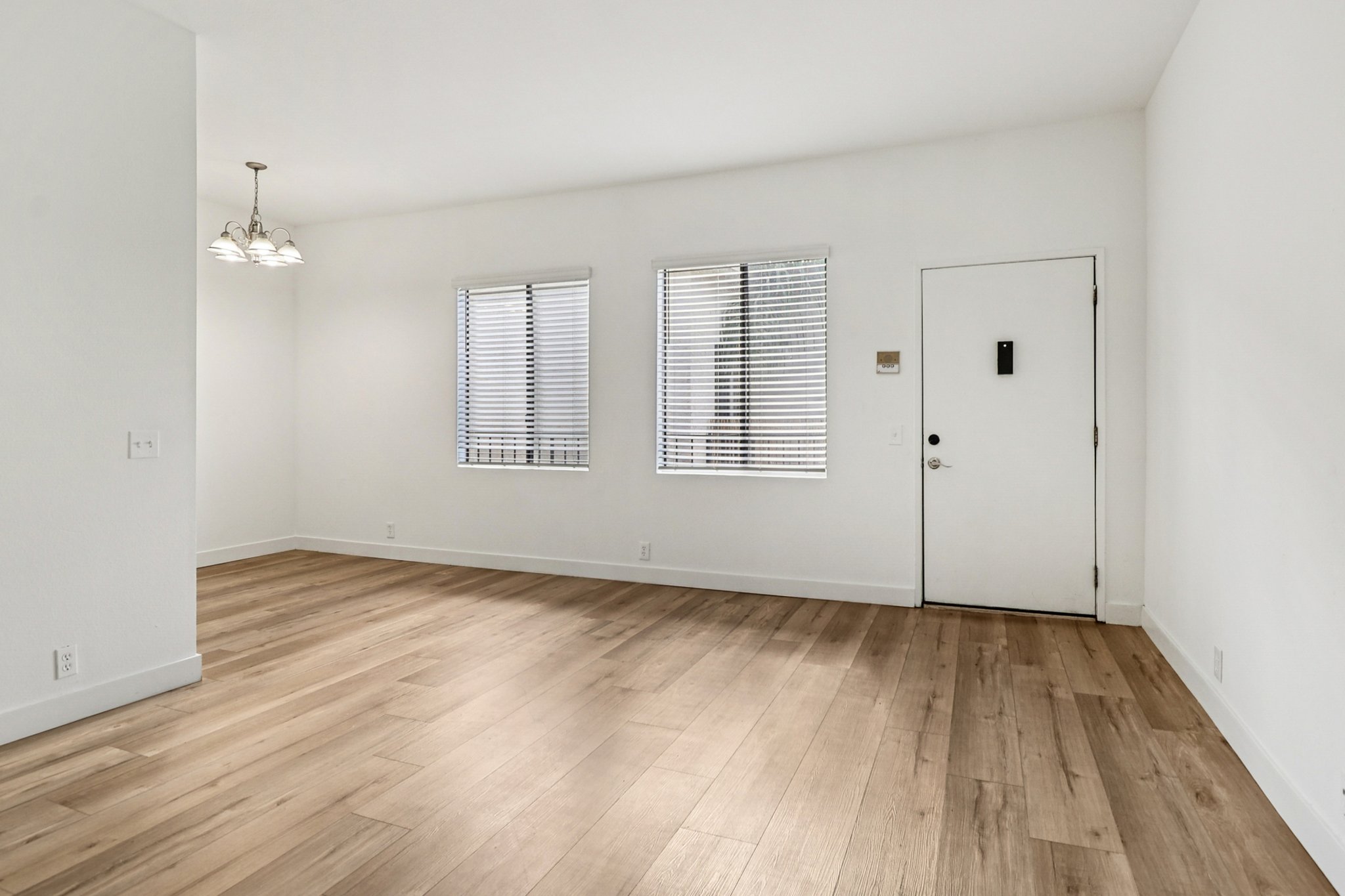 A bright, empty room featuring light-colored walls and wooden flooring. There are two windows with blinds allowing natural light, and a door with a peephole. A chandelier hangs from the ceiling, and the space is clean and uncluttered, suitable for various uses.