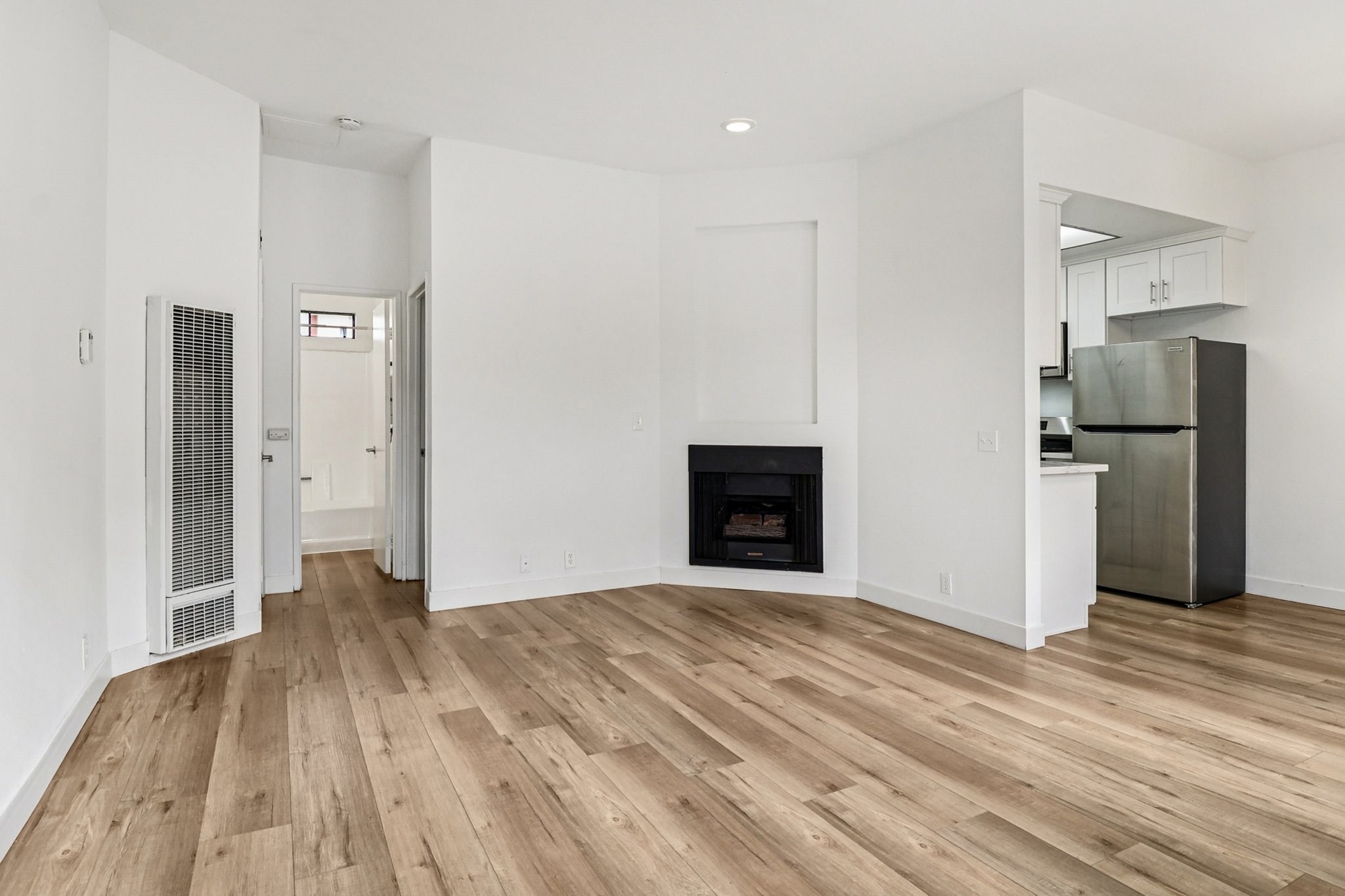 A spacious, modern living room with light wood flooring, featuring a black fireplace. The room is painted white, has large windows, and opens into a kitchen area with a refrigerator visible. Light enters from the left side, and a door leads to another room on the right.