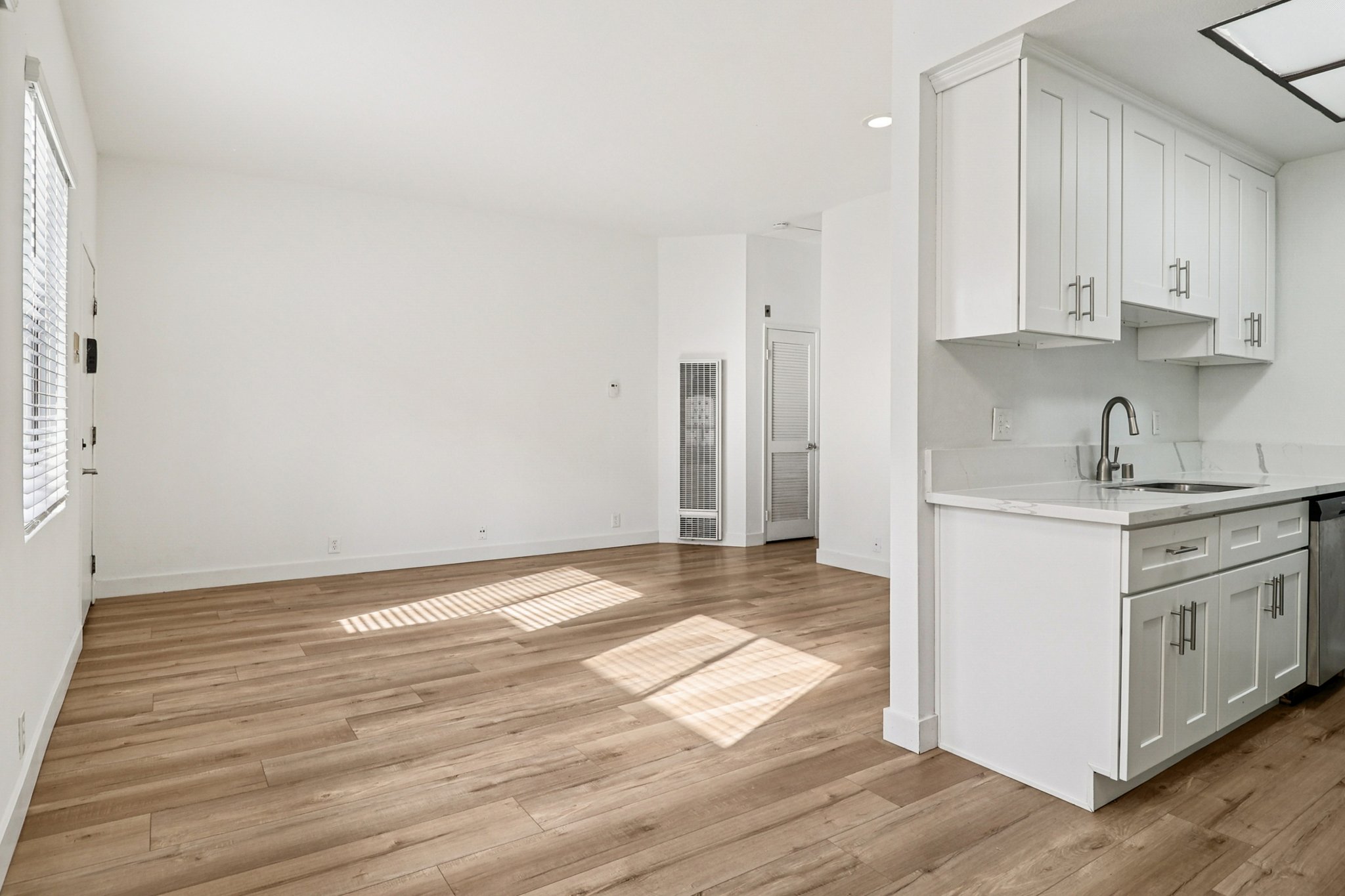 Bright and airy interior of a modern kitchen and living area. The space features light wood flooring, white walls, and large windows allowing natural light to fill the room. The kitchen includes white cabinetry and stainless steel appliances, with a clear division between the kitchen and living area.