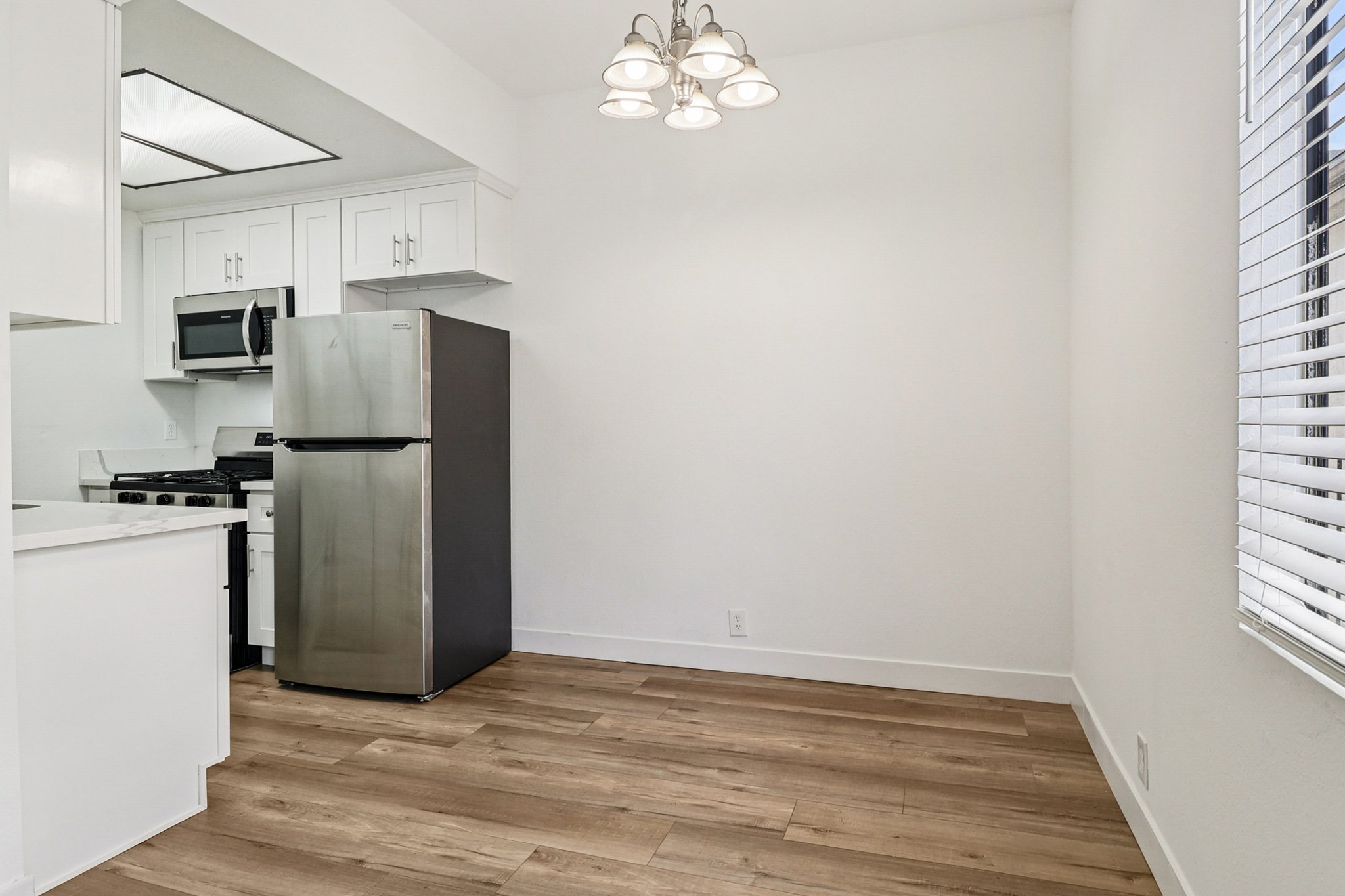 Interior view of a modern kitchen with white cabinets, a stainless steel refrigerator, and a gas stove. The space features light wood flooring, white walls, and a window with blinds. A contemporary light fixture hangs from the ceiling, providing bright illumination. The overall design is clean and minimalistic.