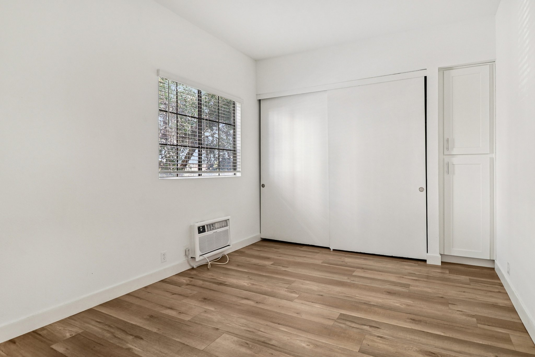 A minimalist room featuring light wooden flooring, a large window with horizontal blinds, and white walls. There is a closet with sliding doors on one side and an air conditioning unit on the wall. The space is empty, creating a bright and open atmosphere.