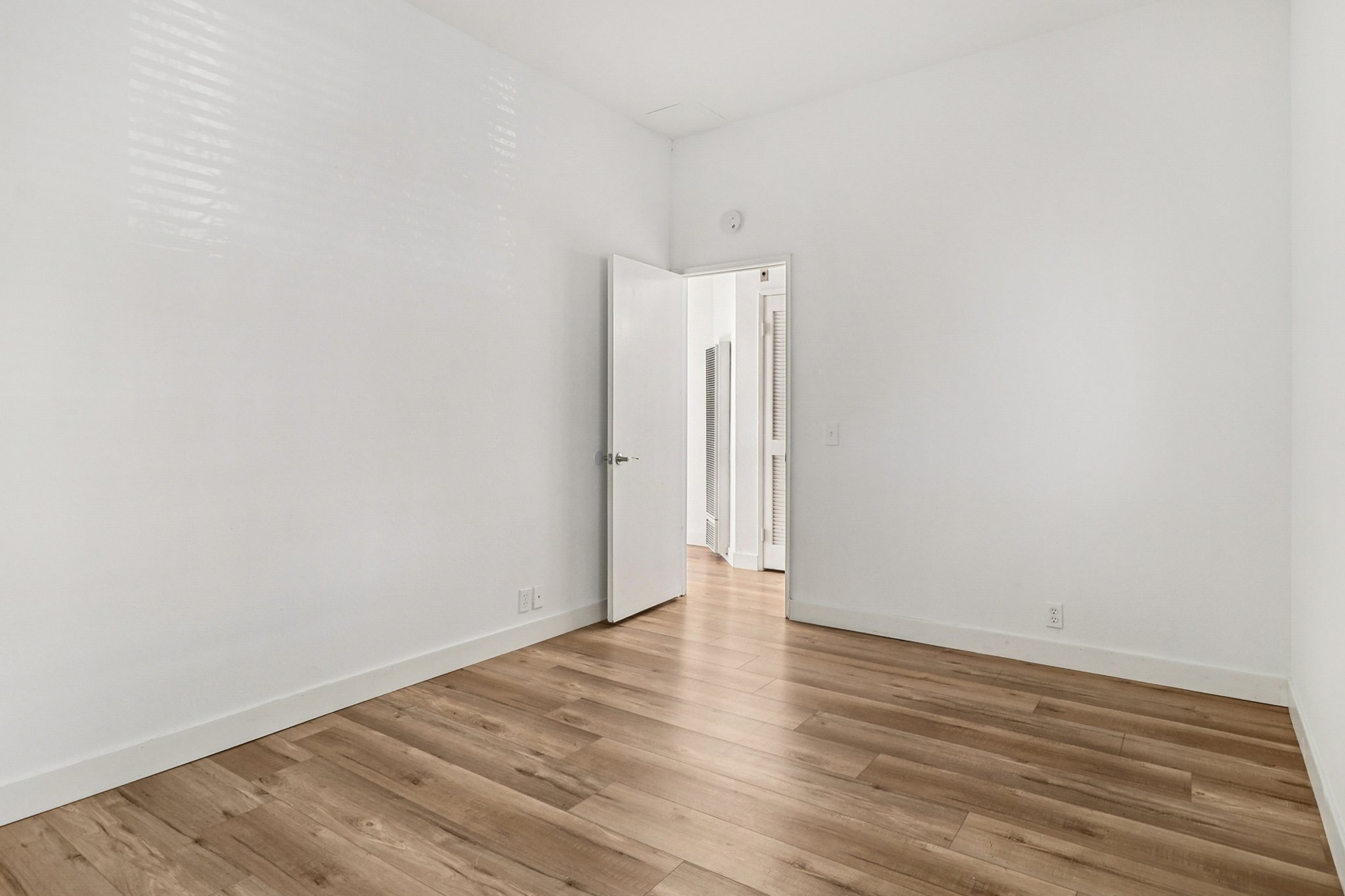 A bright, empty room with white walls and a wooden floor. There is an open door leading to a hallway, and natural light is coming through a window, creating a spacious and clean atmosphere.