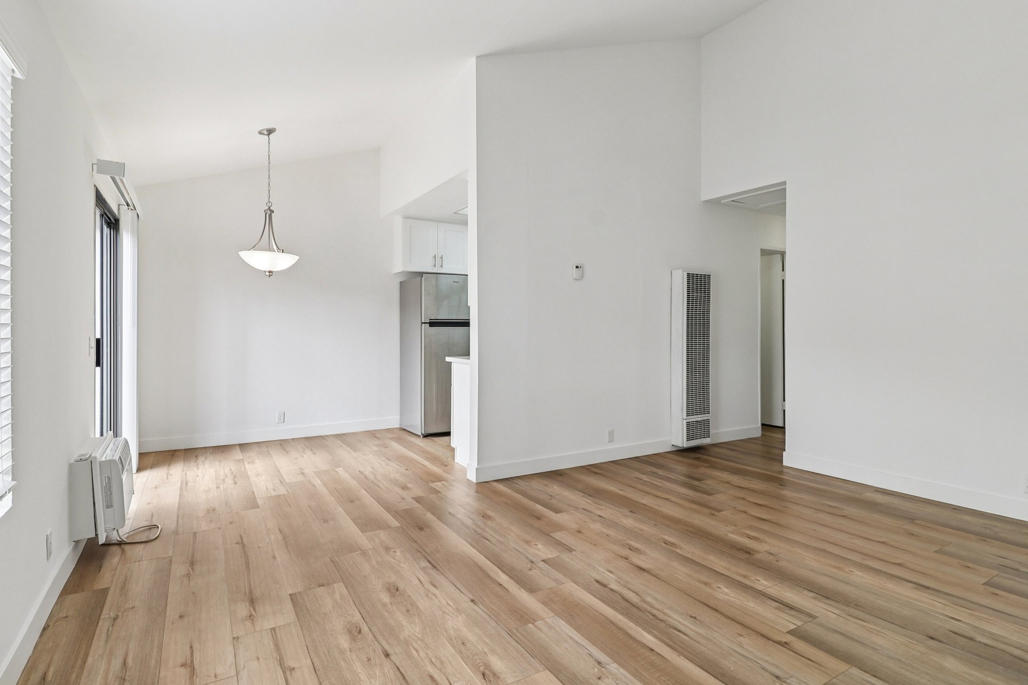 Spacious, modern interior of an empty room featuring wooden flooring, white walls, and large windows. A pendant light hangs from the ceiling, and the kitchen area is visible with stainless steel appliances. The design emphasizes a bright and open layout.