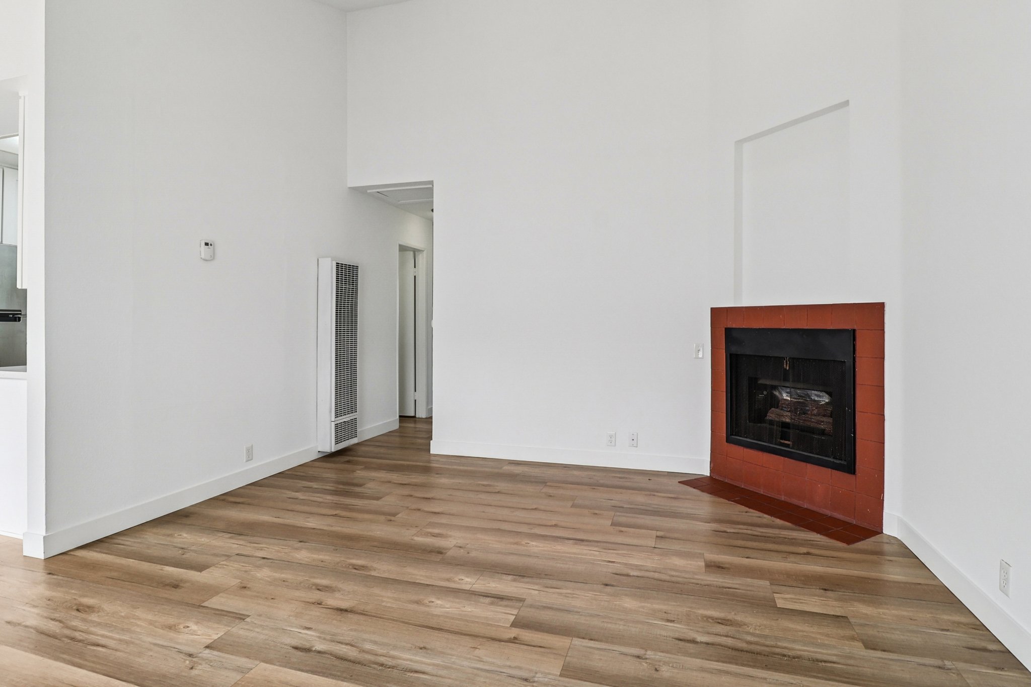 A minimalist interior of a room showcasing light-colored walls and wooden flooring. There is a red-bricked fireplace on one wall, accompanied by a tall radiator. A doorway leads to another space, and the room is brightly lit, creating an open and airy atmosphere.