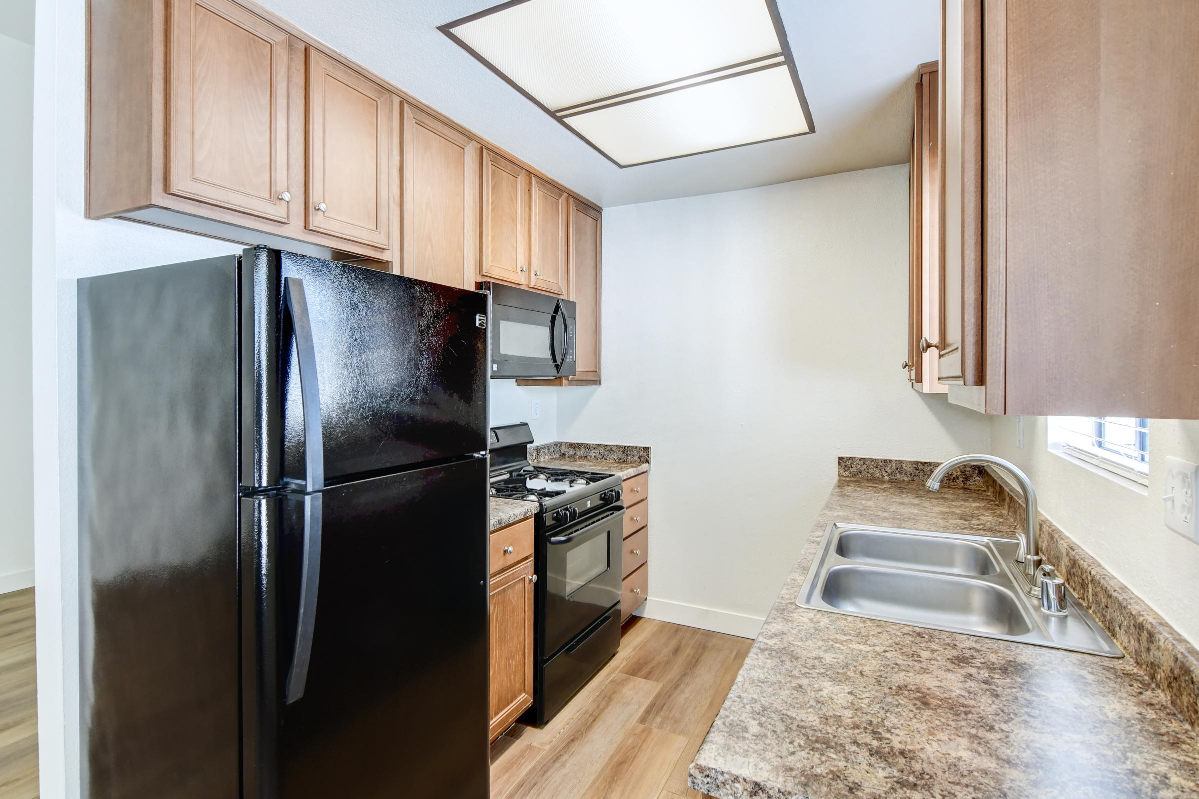 A modern kitchen featuring wooden cabinets, a black refrigerator, a gas stove with an oven, and a stainless steel sink. The countertops are made of a speckled material, and there is a window providing natural light. The floor is finished with light-colored wood laminate.