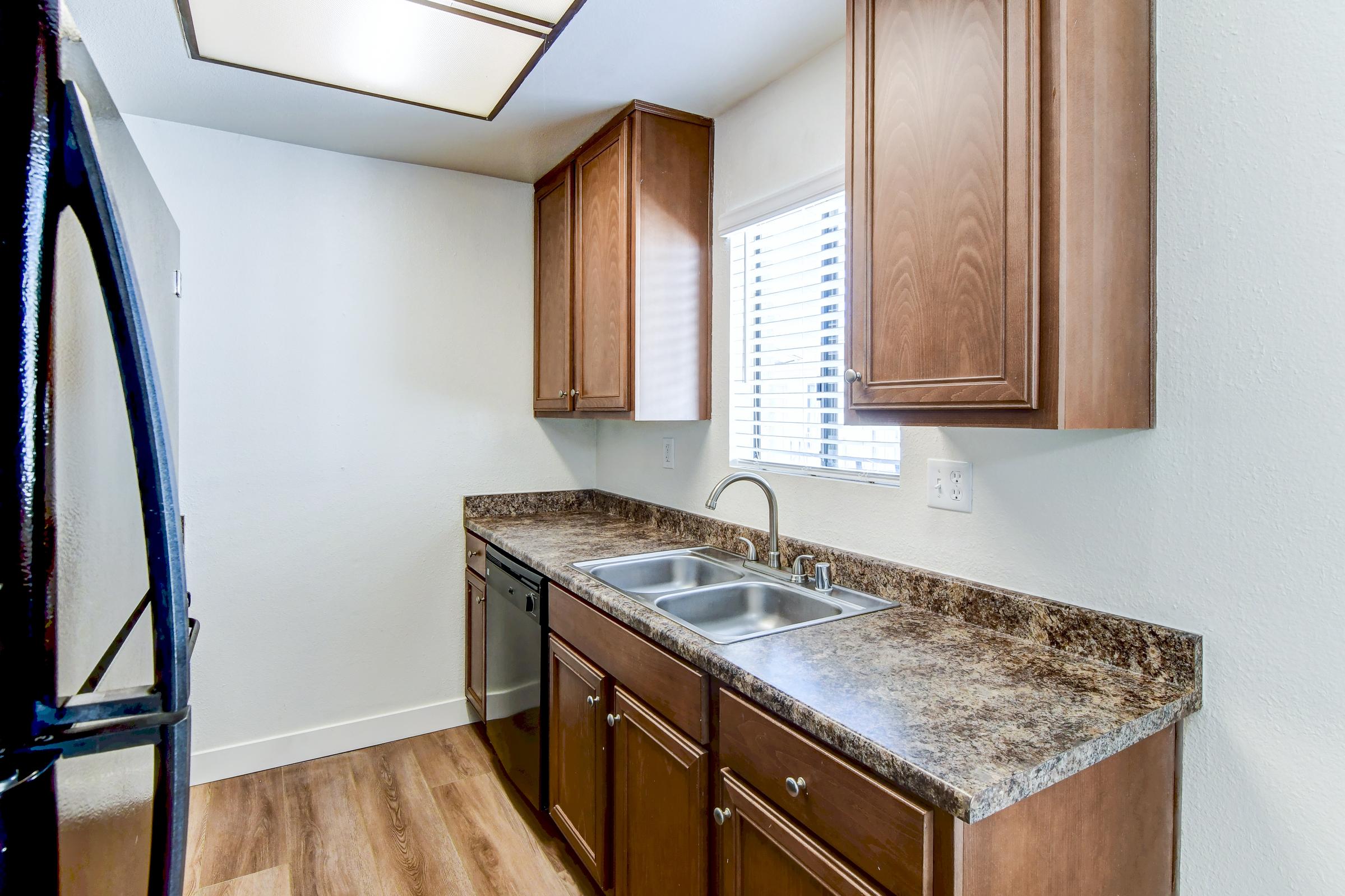 Modern kitchen featuring wooden cabinets, a double sink, and a window with blinds. Black appliances, including a refrigerator and dishwasher, complement the granite countertop. The floor is finished with light wood, creating a bright and inviting atmosphere.