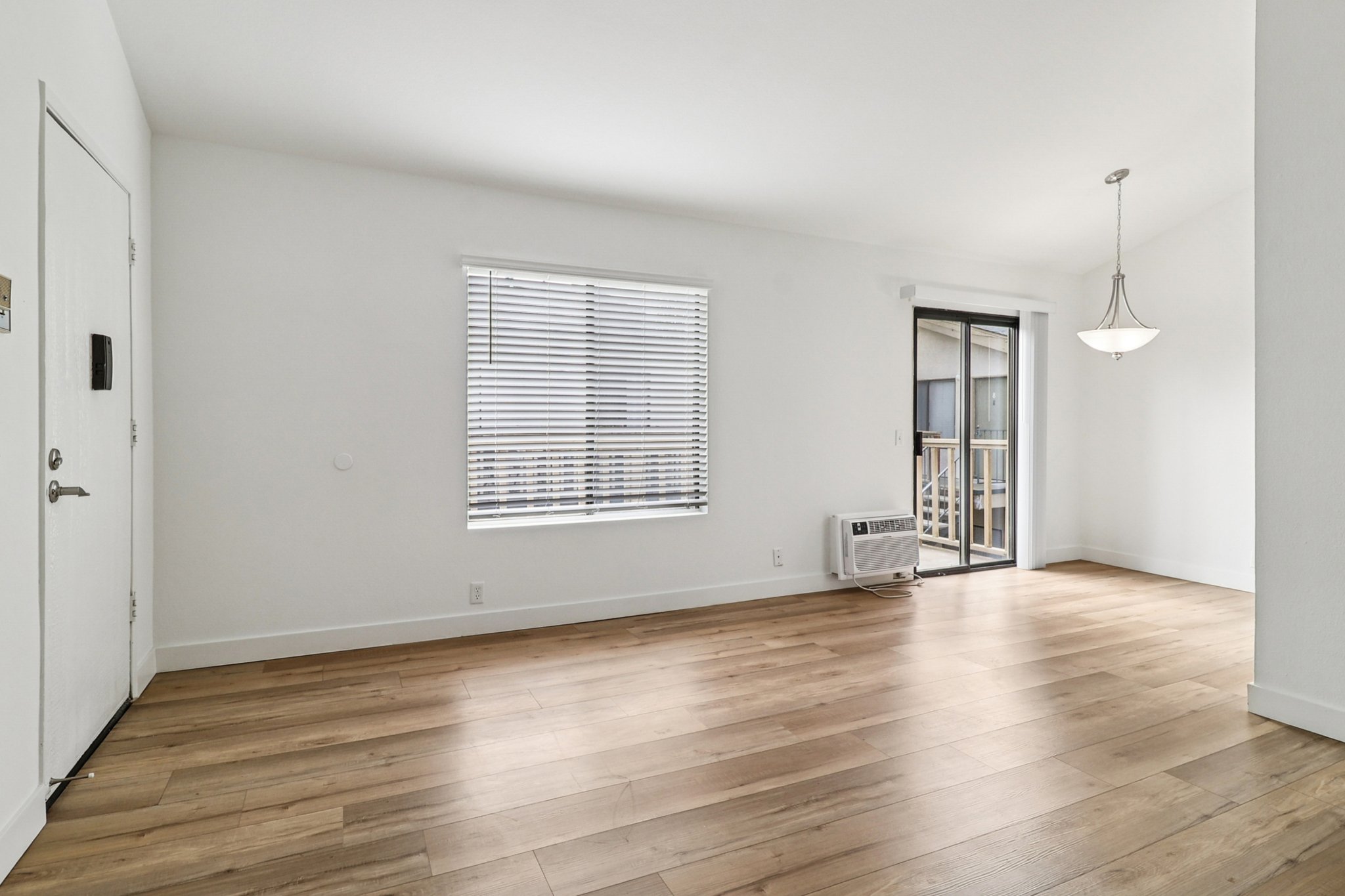 A spacious, empty room with light hardwood flooring and white walls. Large windows with blinds let in natural light, and there's a sliding door leading to a balcony. A light fixture hangs from the ceiling, and an air conditioning unit is visible near the entrance.
