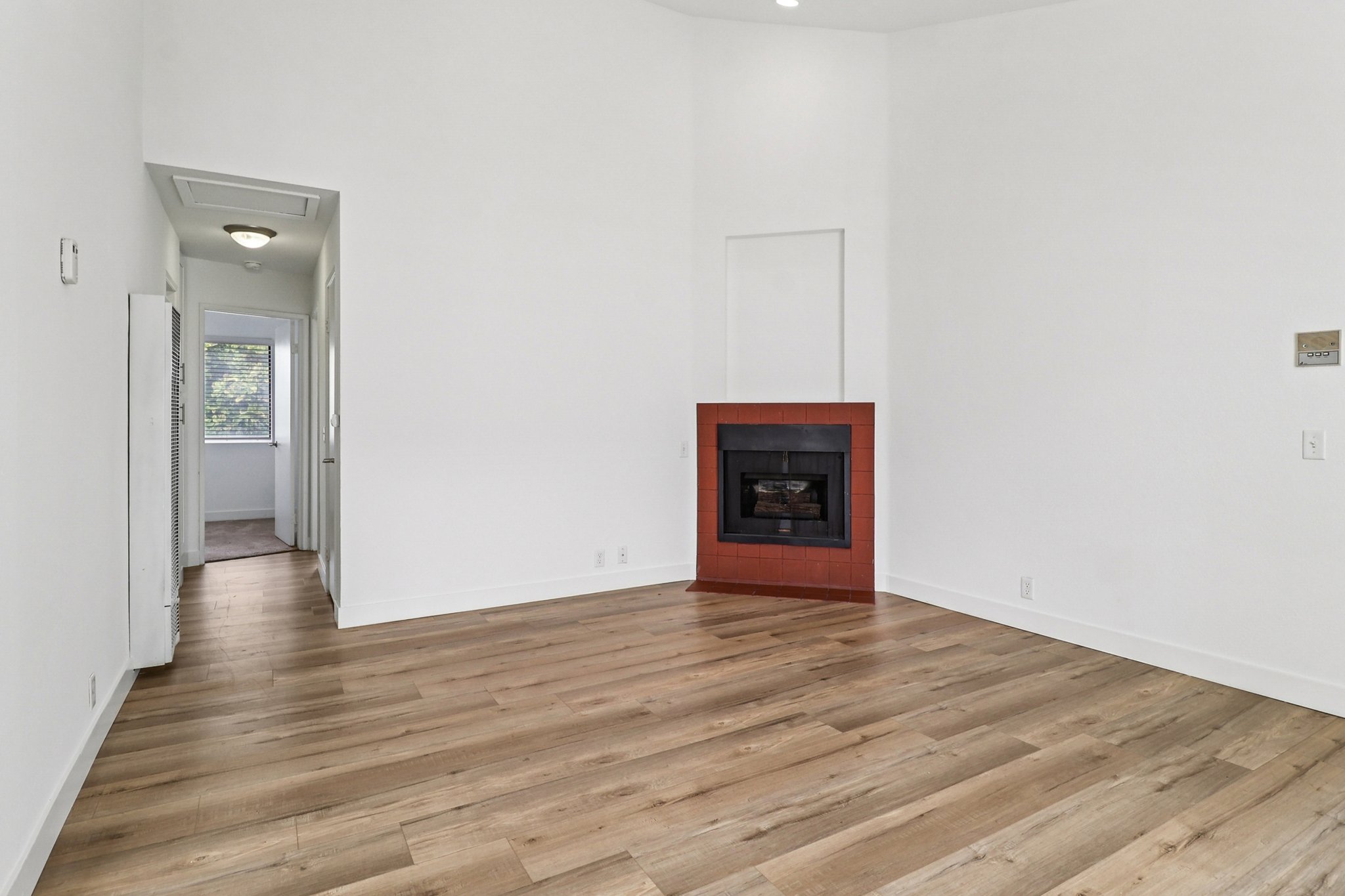 Empty room with light-colored walls and hardwood flooring. A red-brick fireplace is positioned on one wall, creating a focal point. A doorway leads to another room, and a light fixture is visible on the ceiling, adding brightness to the space. Natural light filters in through a window.