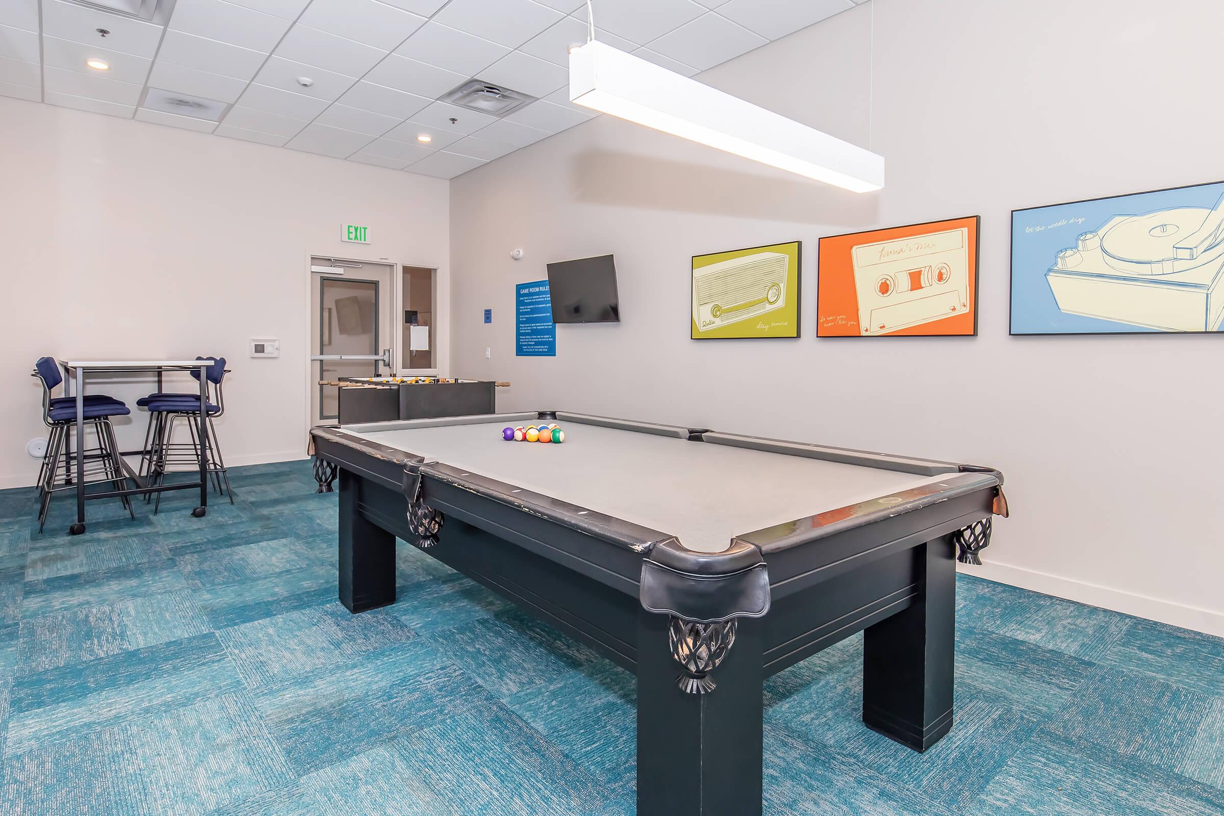 A well-lit recreation room featuring a pool table with colorful balls on it. The walls are decorated with framed art, and there is a seating area in the background. The flooring has a blue carpet, and there's a television mounted on the wall, enhancing the inviting atmosphere for leisure and socializing.