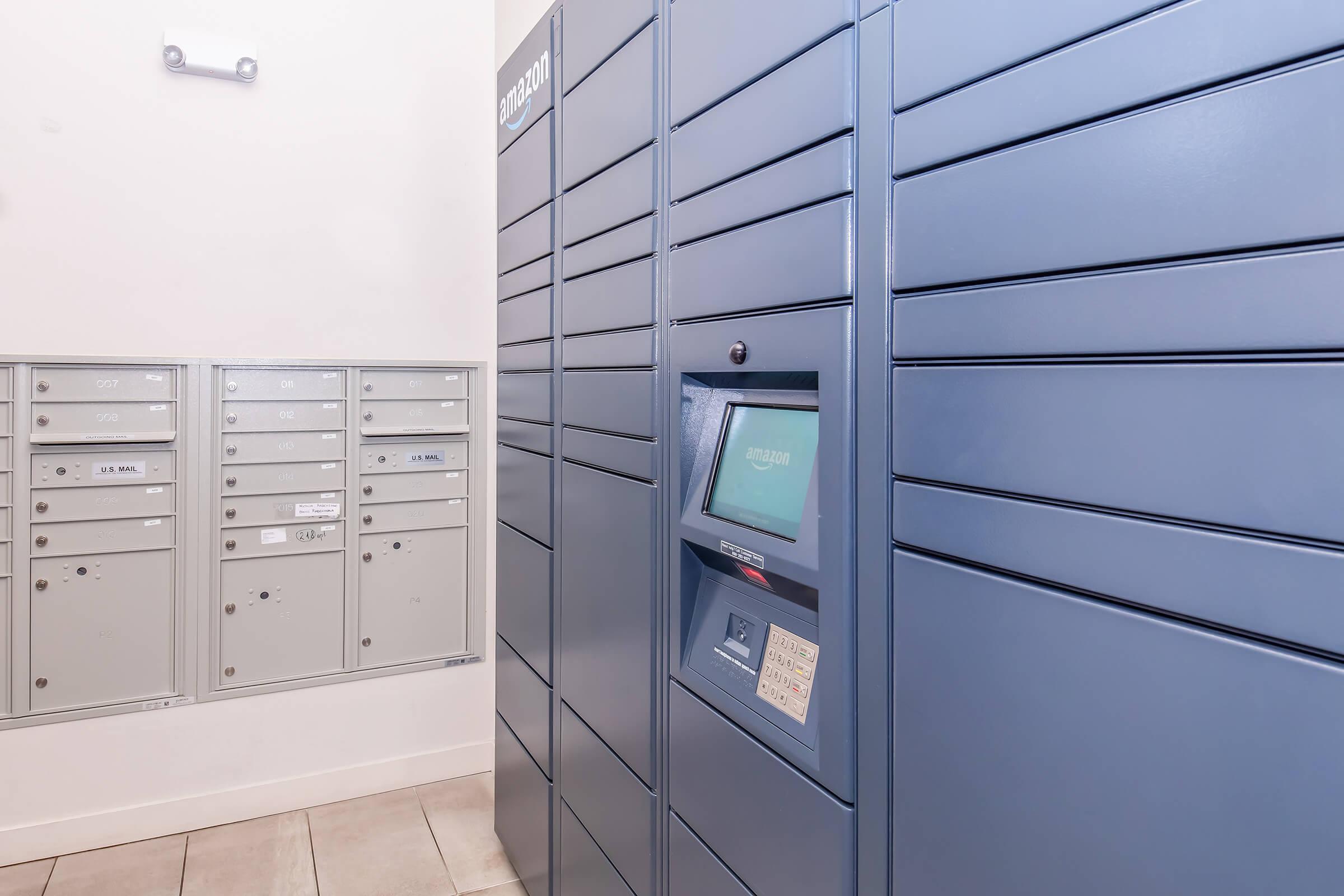 A modern package delivery locker system in a hallway, with blue lockers on the right and gray mailboxes on the left. The lockers have a digital screen for accessing packages, and the environment appears clean and well-lit.