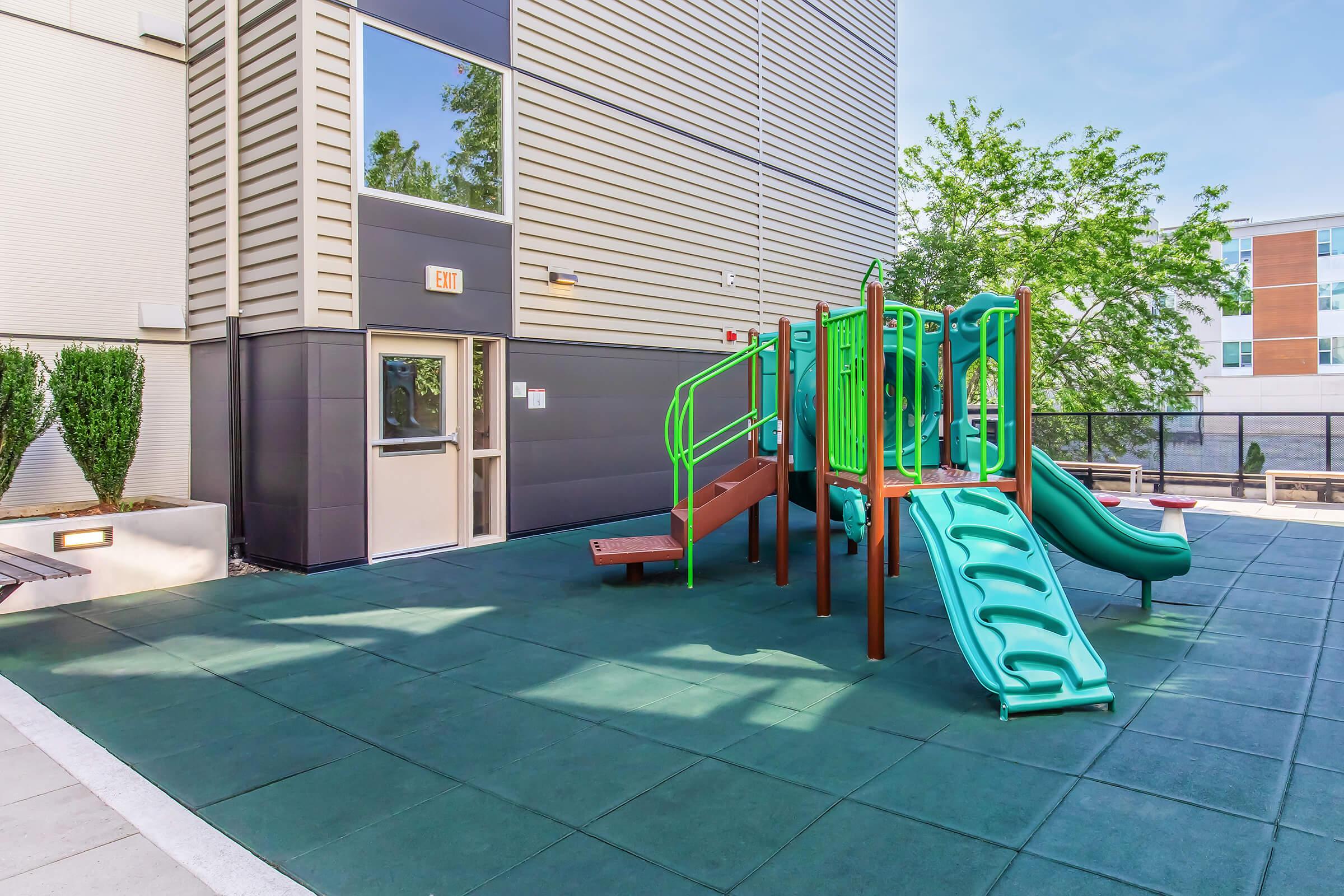 Playground area featuring green slides and climbing structures on padded flooring, adjacent to a building. The setting includes trees and a clear sky, providing a bright and inviting space for children to play.