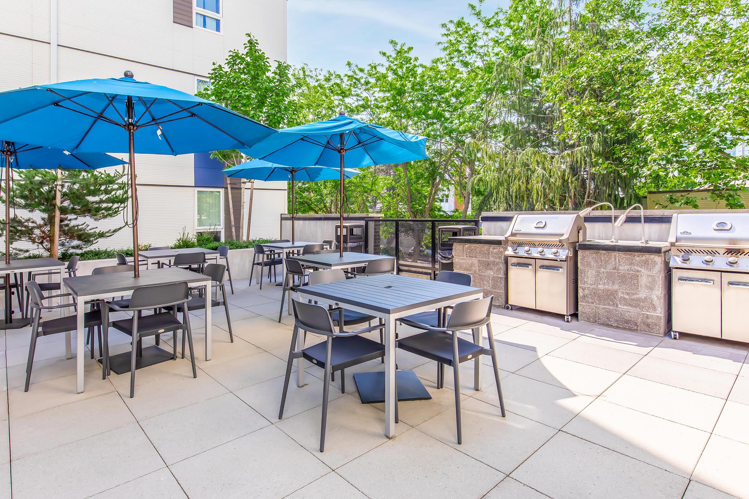 Outdoor patio area featuring several gray tables and chairs under bright blue umbrellas. Two large barbecue grills are positioned against a stone wall, surrounded by greenery. The space is well-lit with a clear blue sky visible overhead, creating a welcoming atmosphere for dining and gatherings.
