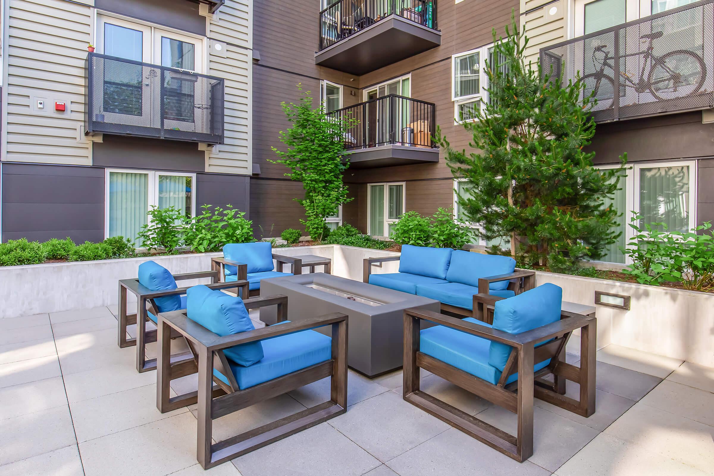 A modern outdoor lounge area featuring a stylish set of blue cushioned chairs and a rectangular fire pit table. Surrounding greenery and shrubs add a touch of nature, while nearby balconies offer additional living space. The setting is designed for relaxation and socializing in an urban environment.