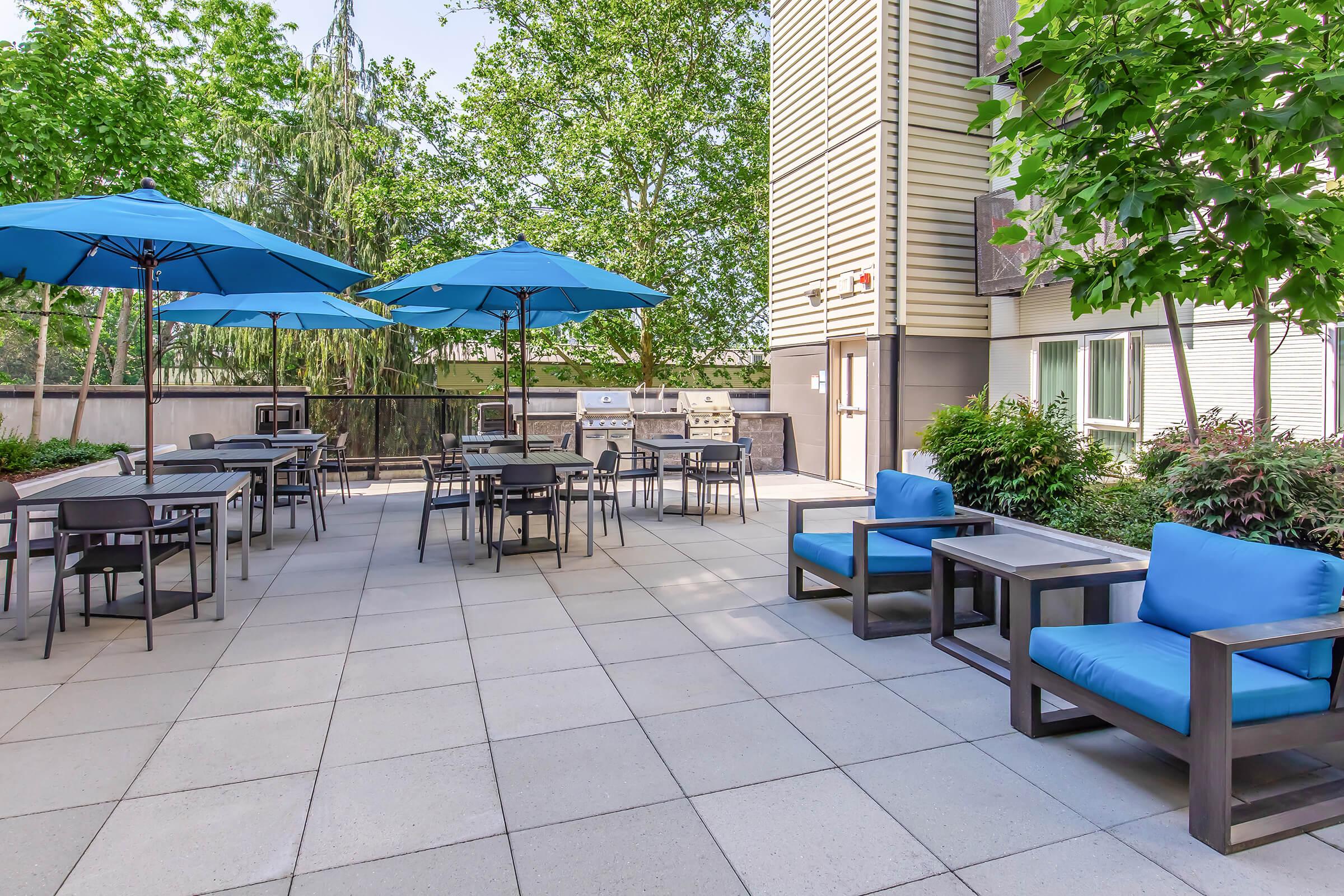 A spacious outdoor patio area featuring several blue umbrellas providing shade over tables and chairs. Comfortable blue lounge seating is arranged on the side, surrounded by greenery and trees, creating a relaxing and inviting atmosphere for gatherings or leisure time.