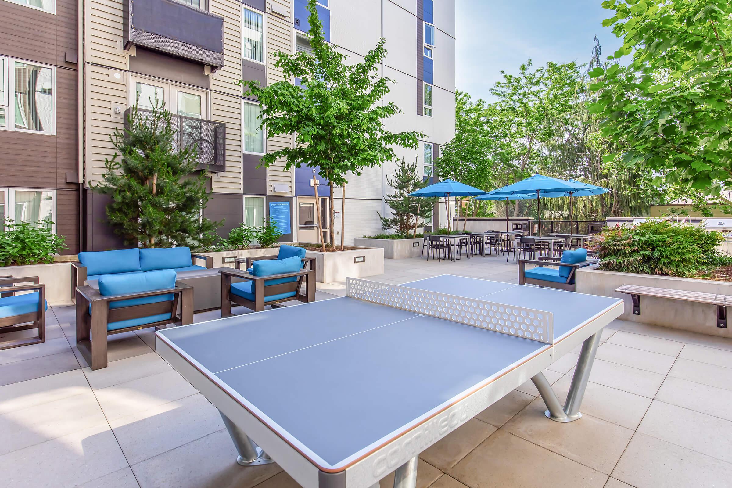 A vibrant outdoor recreational area featuring a ping pong table surrounded by lush greenery, blue lounge chairs, and patio umbrellas. The modern building in the background adds to the inviting atmosphere, providing a perfect spot for relaxation and socializing.