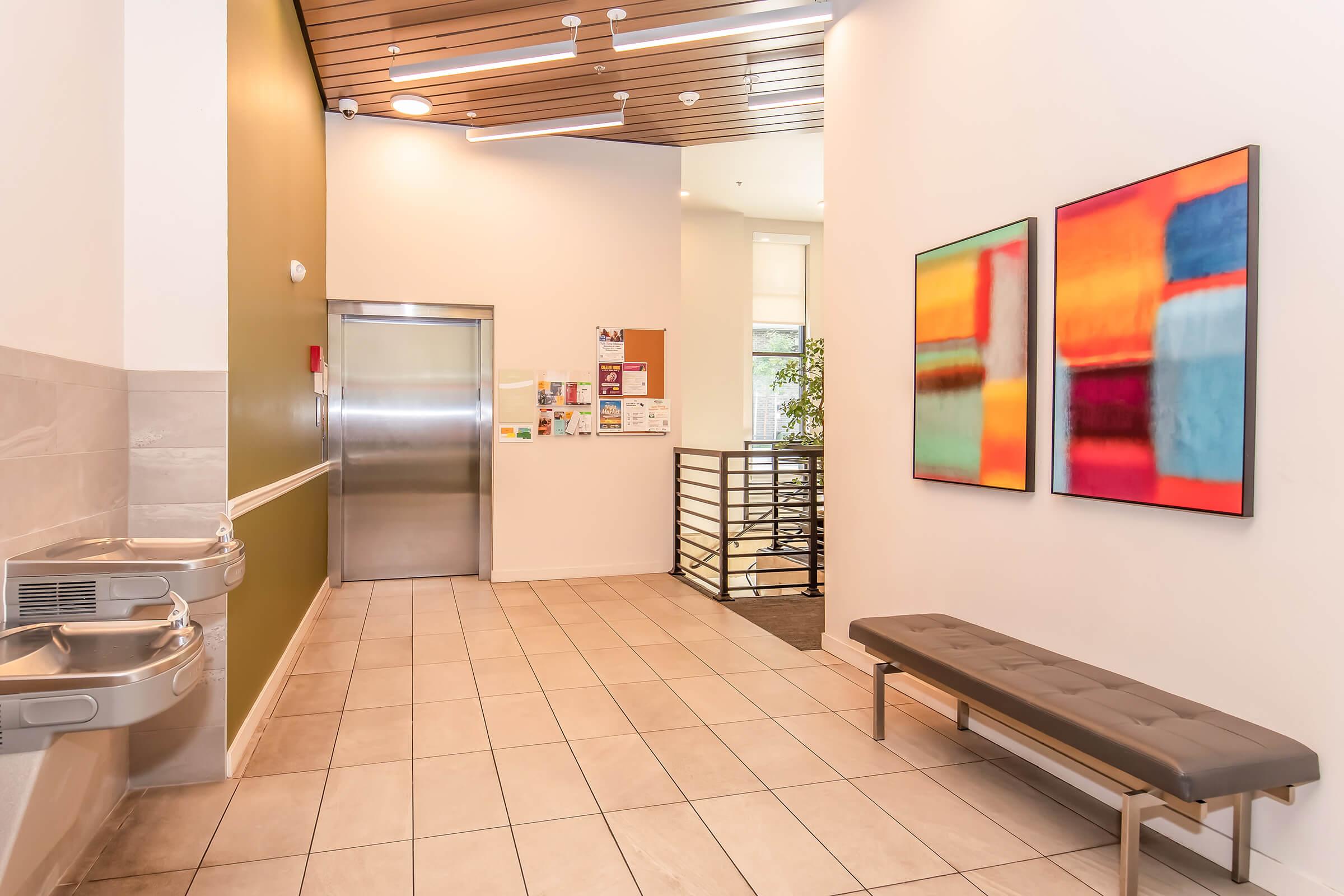 A neatly designed hallway featuring an elevator, two drinking fountains, and a gray bench. On the wall, there are two colorful abstract paintings with vibrant hues. The space has modern lighting and a clean tile floor, contributing to a bright and inviting atmosphere.