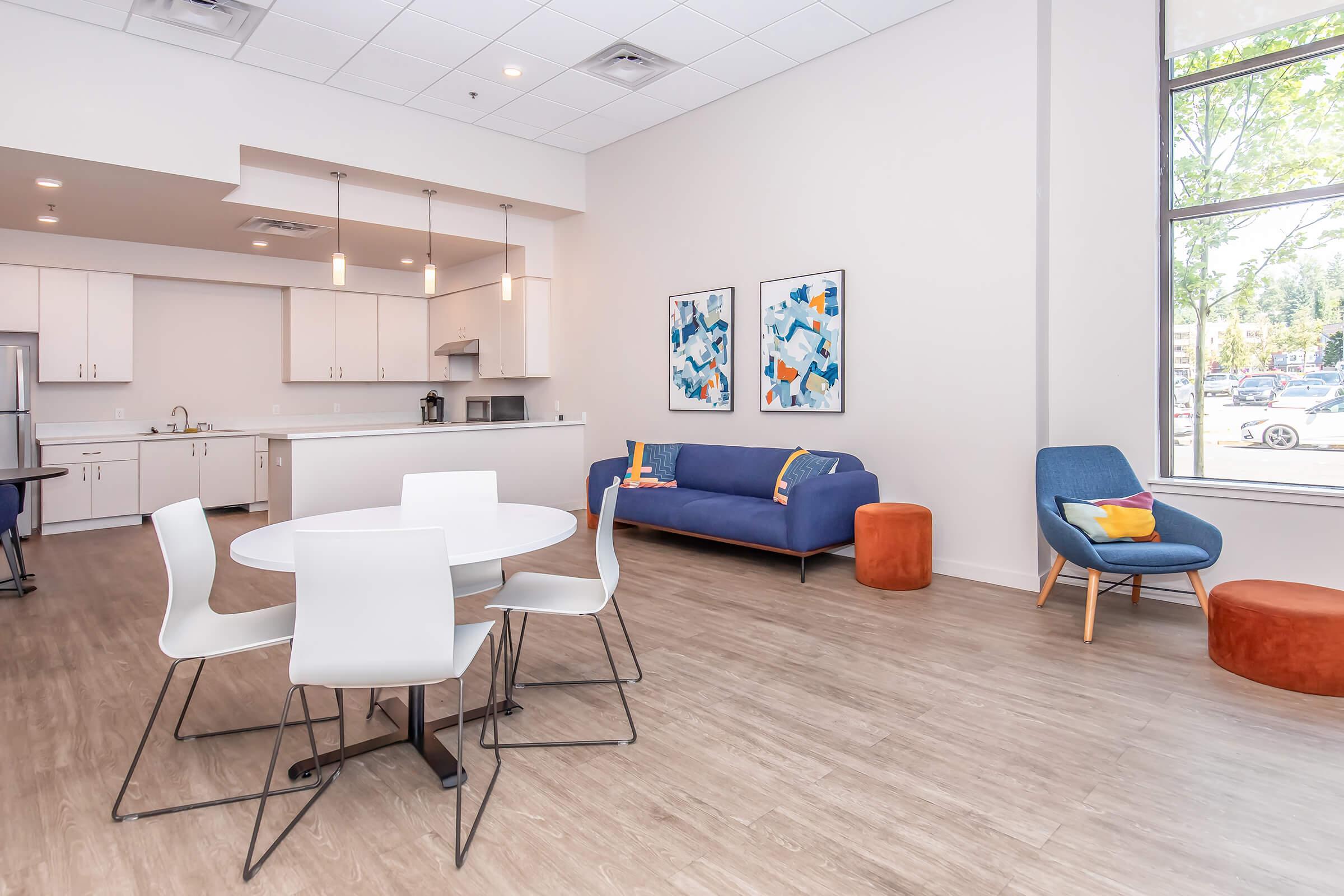 Modern open-concept living space featuring a kitchen area with white cabinetry, a round dining table with white chairs, a blue sofa, and colorful accent pillows. A cozy orange pouf and a stylish armchair add warmth. Large windows provide natural light, enhancing the inviting atmosphere.