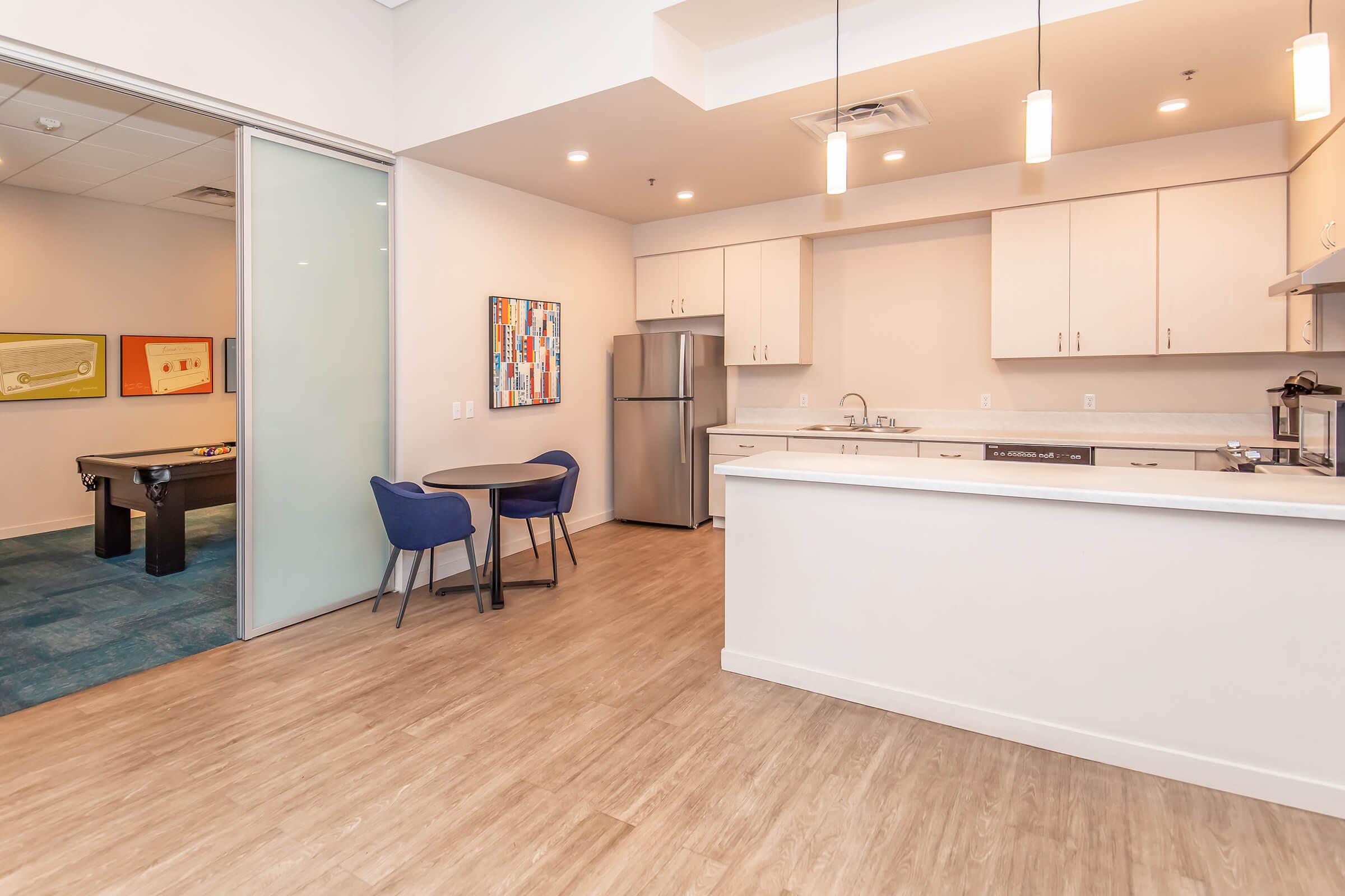 Modern kitchen space featuring sleek white cabinets, a silver refrigerator, and a small round dining table with blue chairs. The area has hardwood-style flooring and pendant lighting. A glass partition leads to a room with a pool table, adding to the contemporary atmosphere.