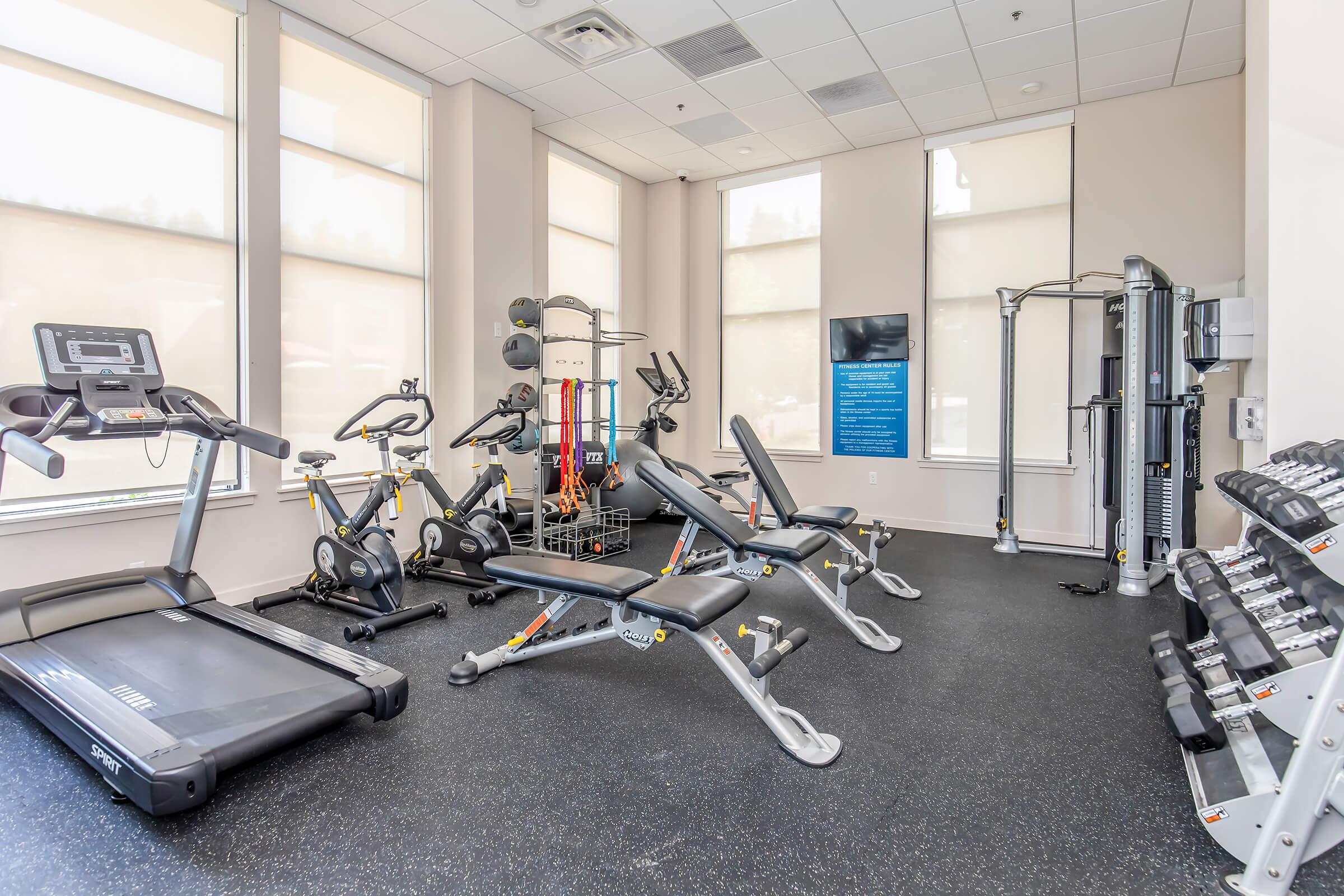 A well-equipped fitness room featuring a treadmill, stationary bike, strength training machines, and a rack of dumbbells. Large windows provide natural light, and a blue informational poster is displayed on the wall. The space has a modern design with a black rubber floor.