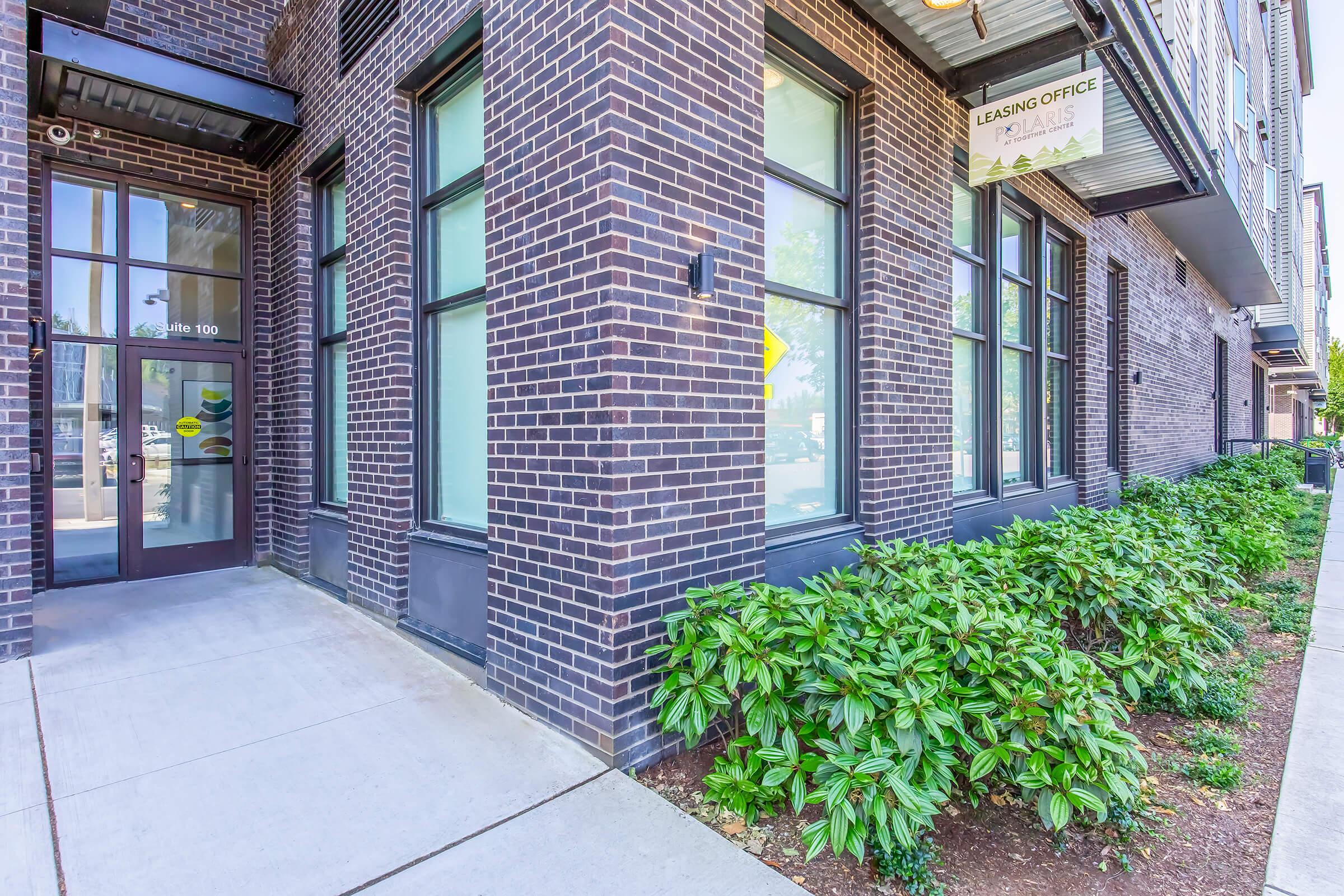 Exterior view of a modern leasing office entrance, featuring large windows and a welcoming door. The brick building has greenery planted along the sidewalk, creating a pleasant urban environment. A sign indicating "Leasing Office" is visible above the entrance.
