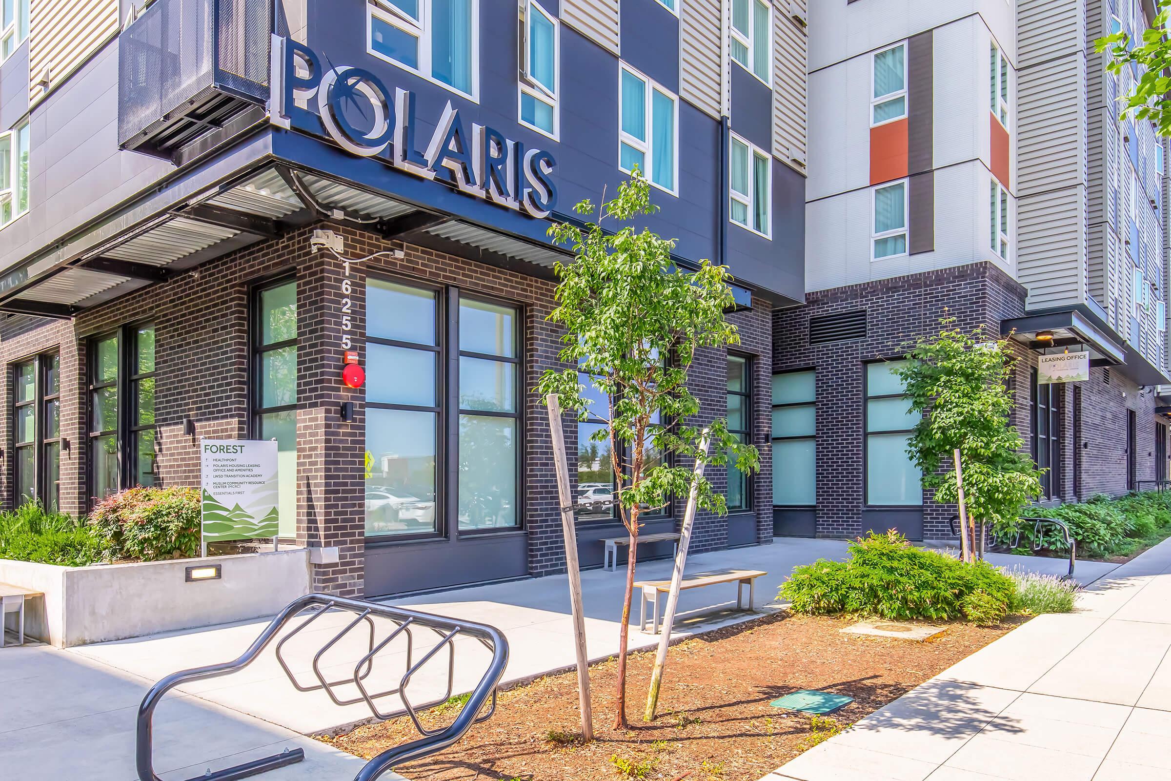 A modern apartment building named "Polaris" featuring sleek architecture, large windows, and landscaping with trees and shrubs. The entrance is designed with a bench and a bike rack, suggesting a welcoming environment for residents and visitors.