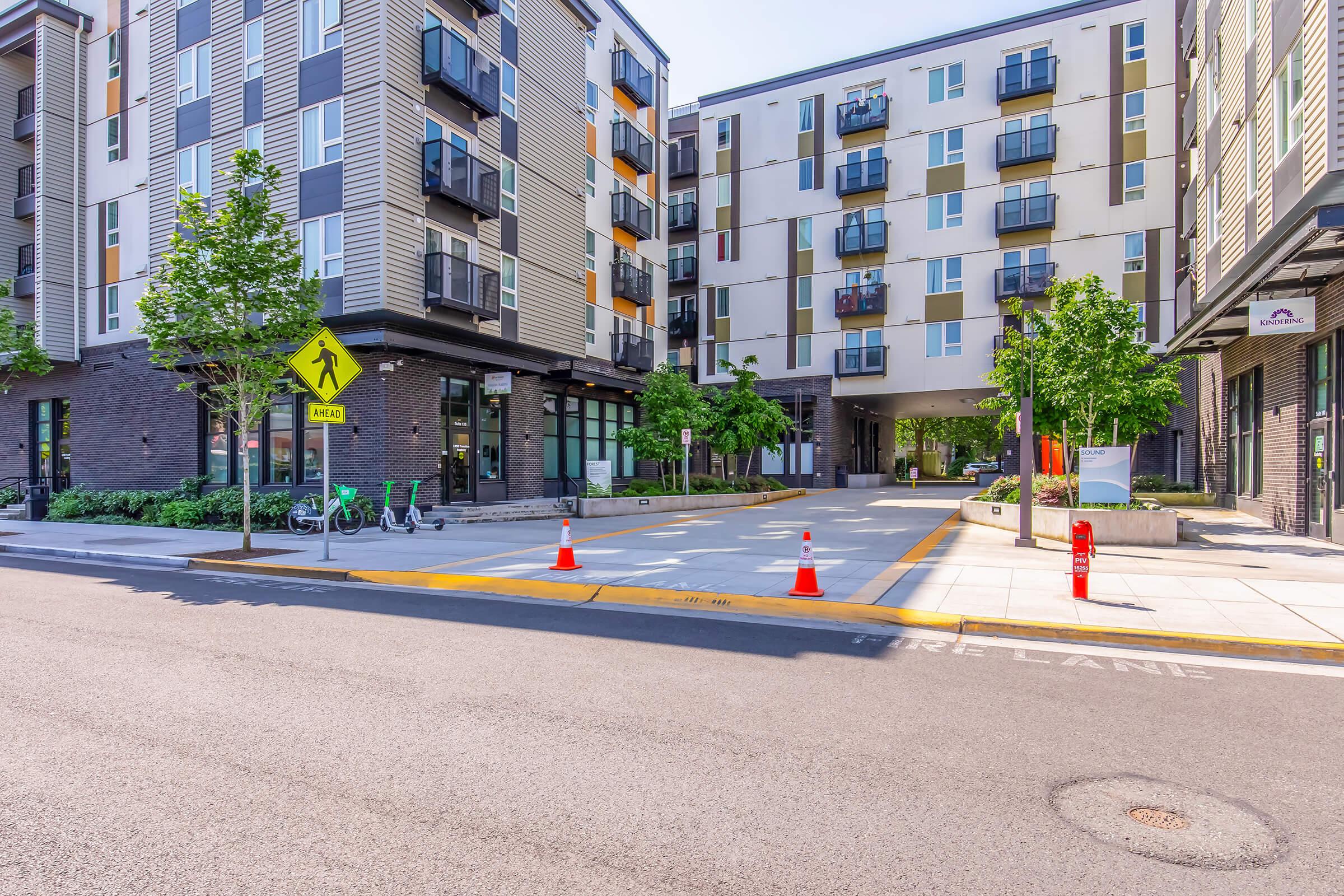 A modern urban building with multiple stories, featuring a mix of balconies and large windows. The area includes a wide sidewalk, trees, and planters. Traffic cones are placed on the street, indicating a pedestrian zone. Bicycles are parked nearby, suggesting a bike-friendly environment.