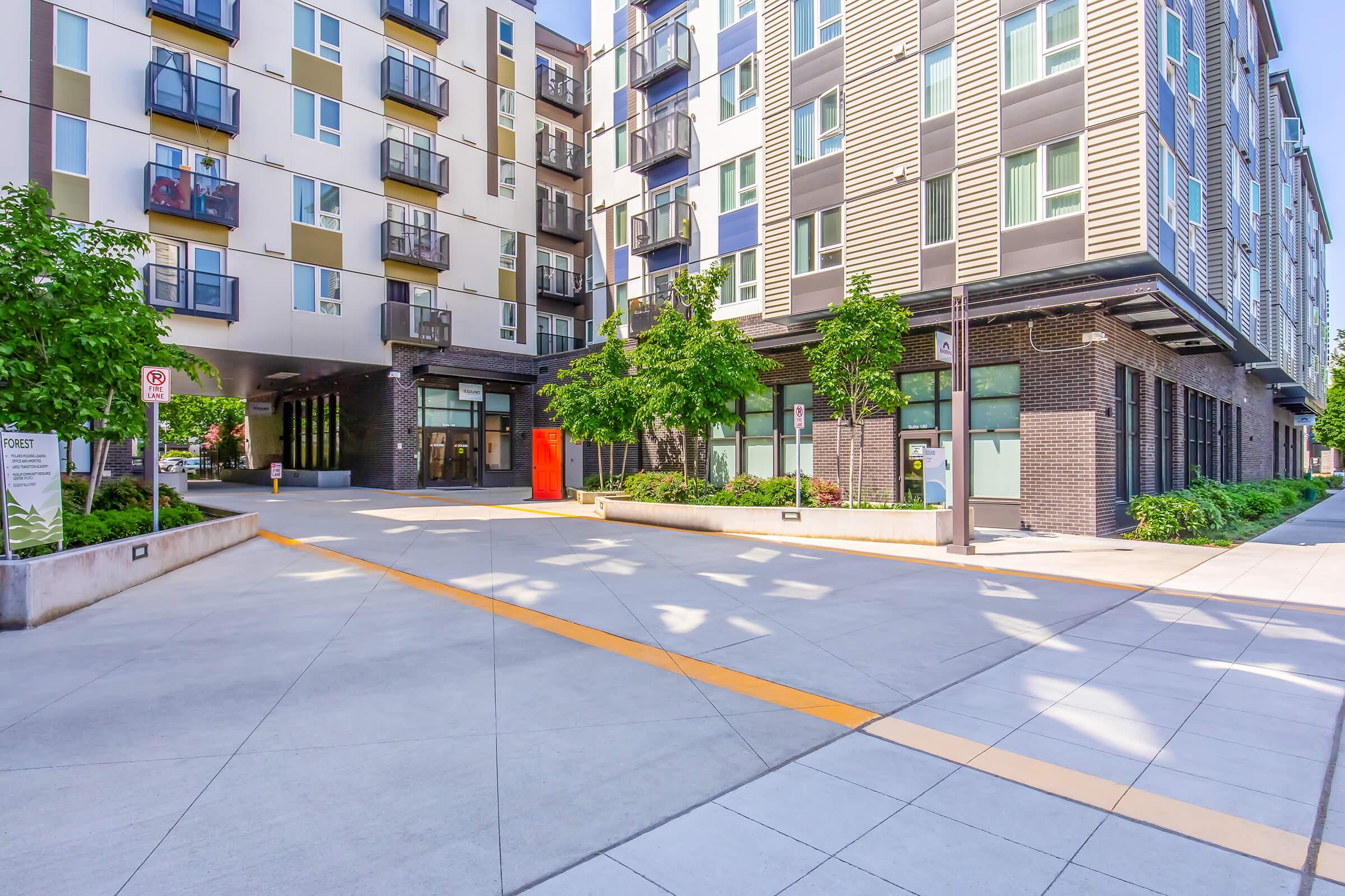 Modern urban apartment building featuring a sleek exterior design with balconies. The image shows a wide, paved pathway lined with small trees and planters, leading to the building's entrance, which is marked by a vibrant orange door. Natural light casts shadows on the pavement, enhancing the contemporary aesthetic.