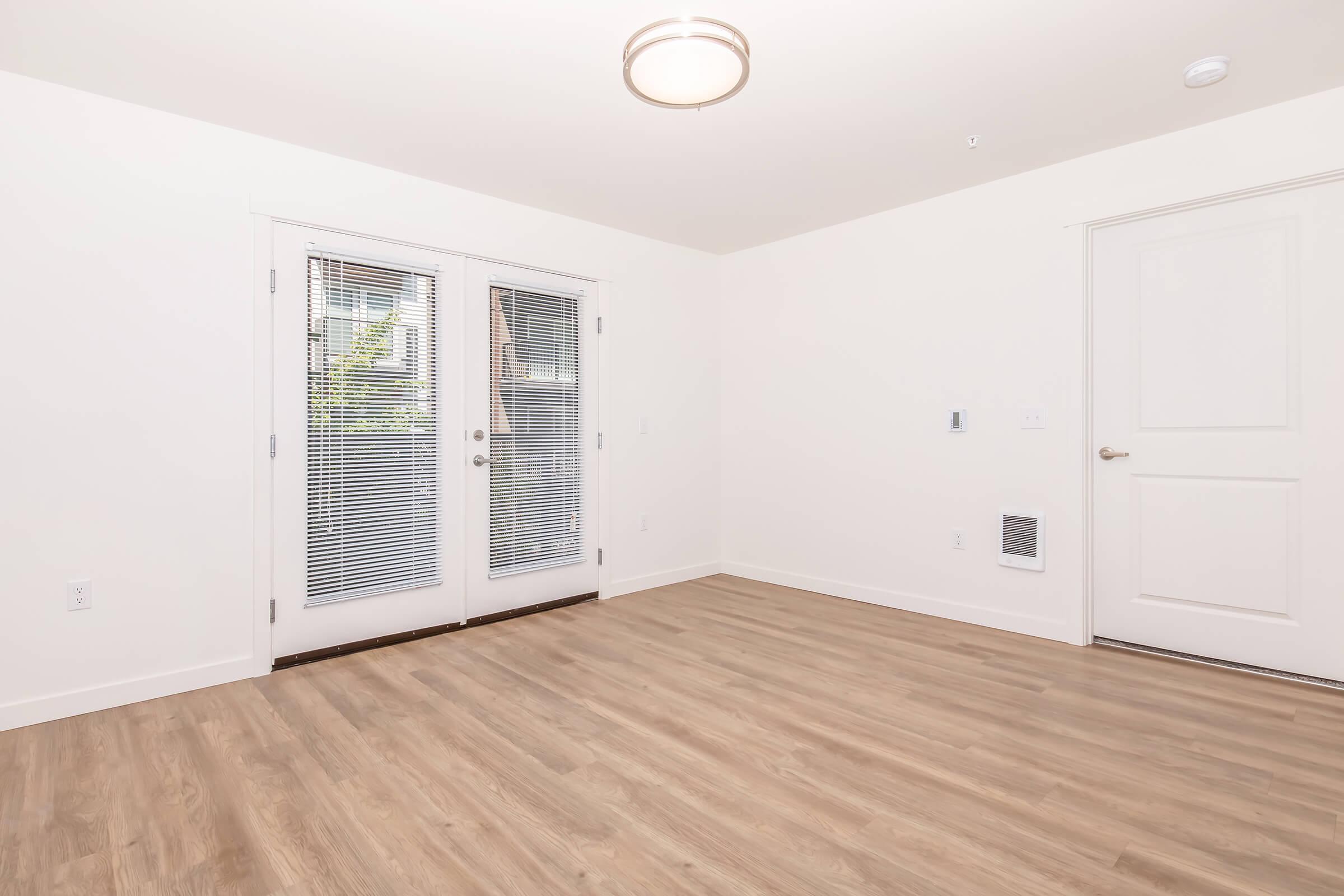 A spacious room with light-colored walls and hardwood flooring. It features double doors with blinds that lead outside, a ceiling light fixture, and a door on the right leading to another room. The space is bright and minimalist, creating an inviting atmosphere.