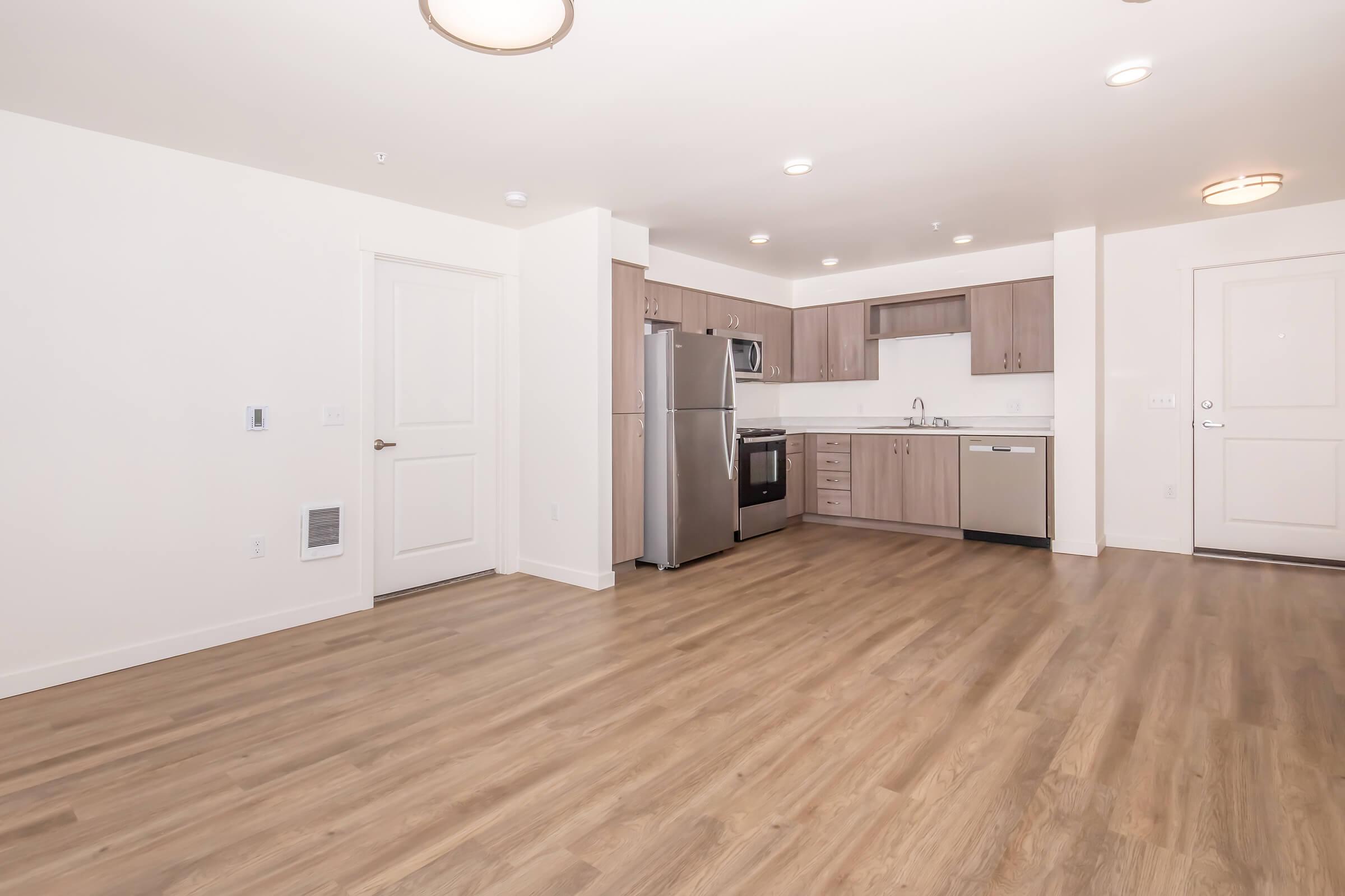 A modern kitchenette with wooden floors, featuring stainless steel appliances including a refrigerator, oven, and dishwasher. Light-colored cabinets and countertops provide a clean aesthetic. To the right, a door leads outside, and there is also a wall with a white door and an air vent.