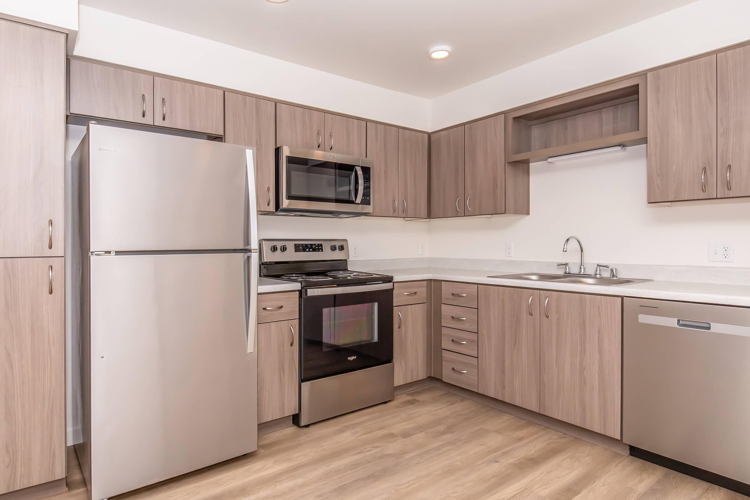 Modern kitchen featuring light wood cabinetry, stainless steel appliances, including a refrigerator, microwave, and oven. The countertop is white with a subtle pattern, and there is a double sink. Natural light fills the space, highlighting the clean and contemporary design.