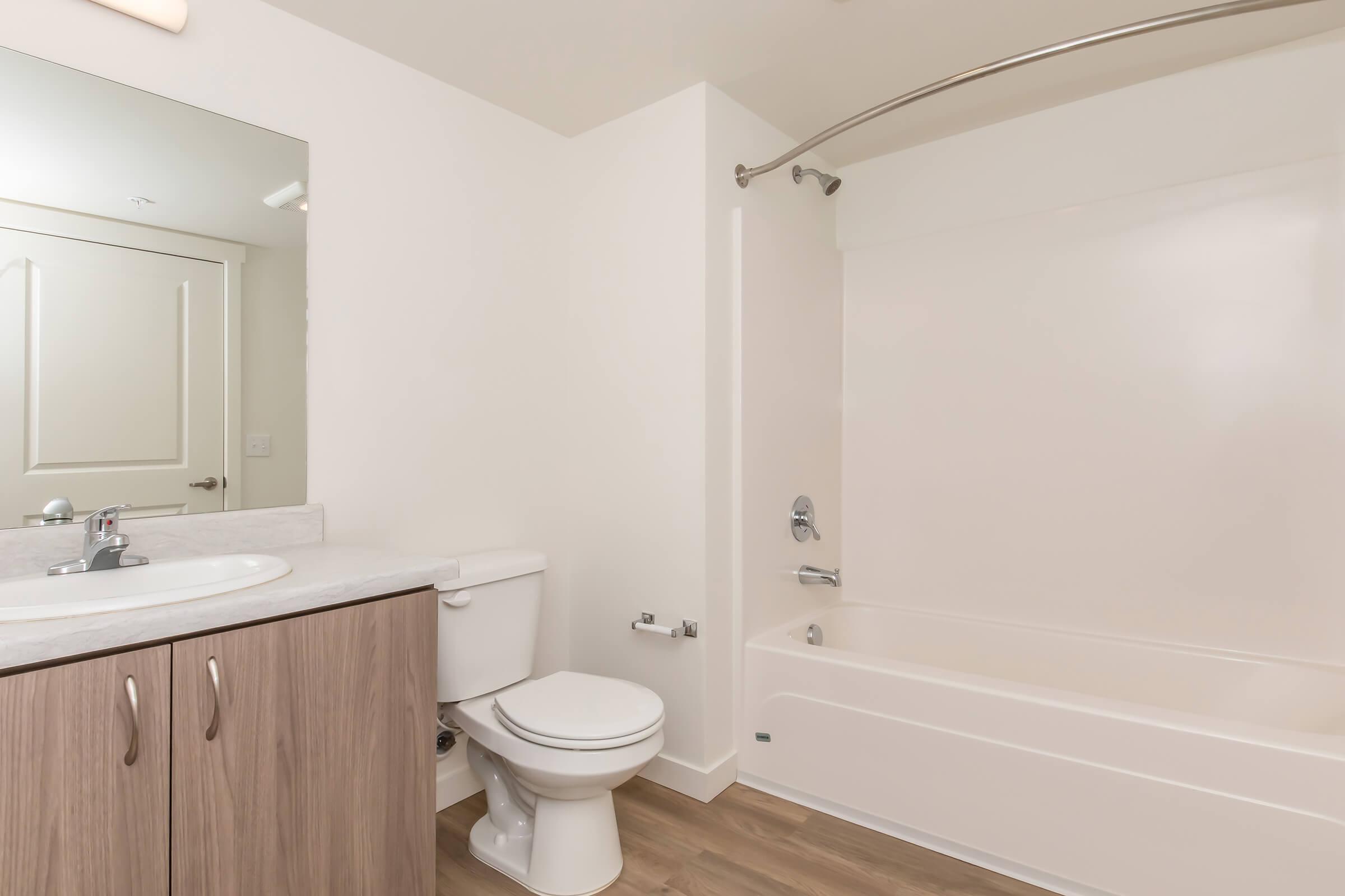 A clean, modern bathroom featuring a white bathtub with a shower, a toilet, and a vanity with a sink. The space includes a large mirror and neutral-colored walls, with wood flooring that adds warmth to the design.