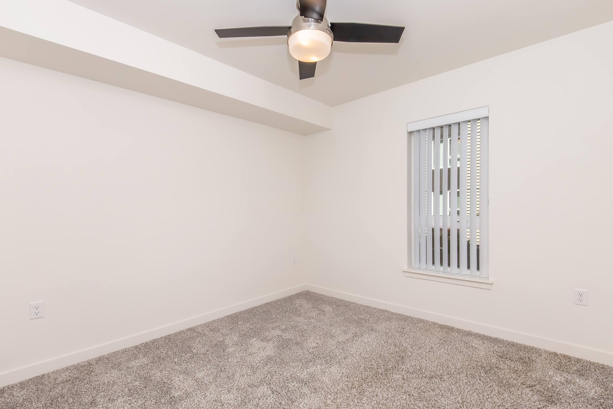 Empty bedroom with light-colored walls, a ceiling fan, a window with vertical blinds, and plush carpet flooring. The room is bright and spacious, featuring neutral tones and minimal furnishings, creating an inviting atmosphere.
