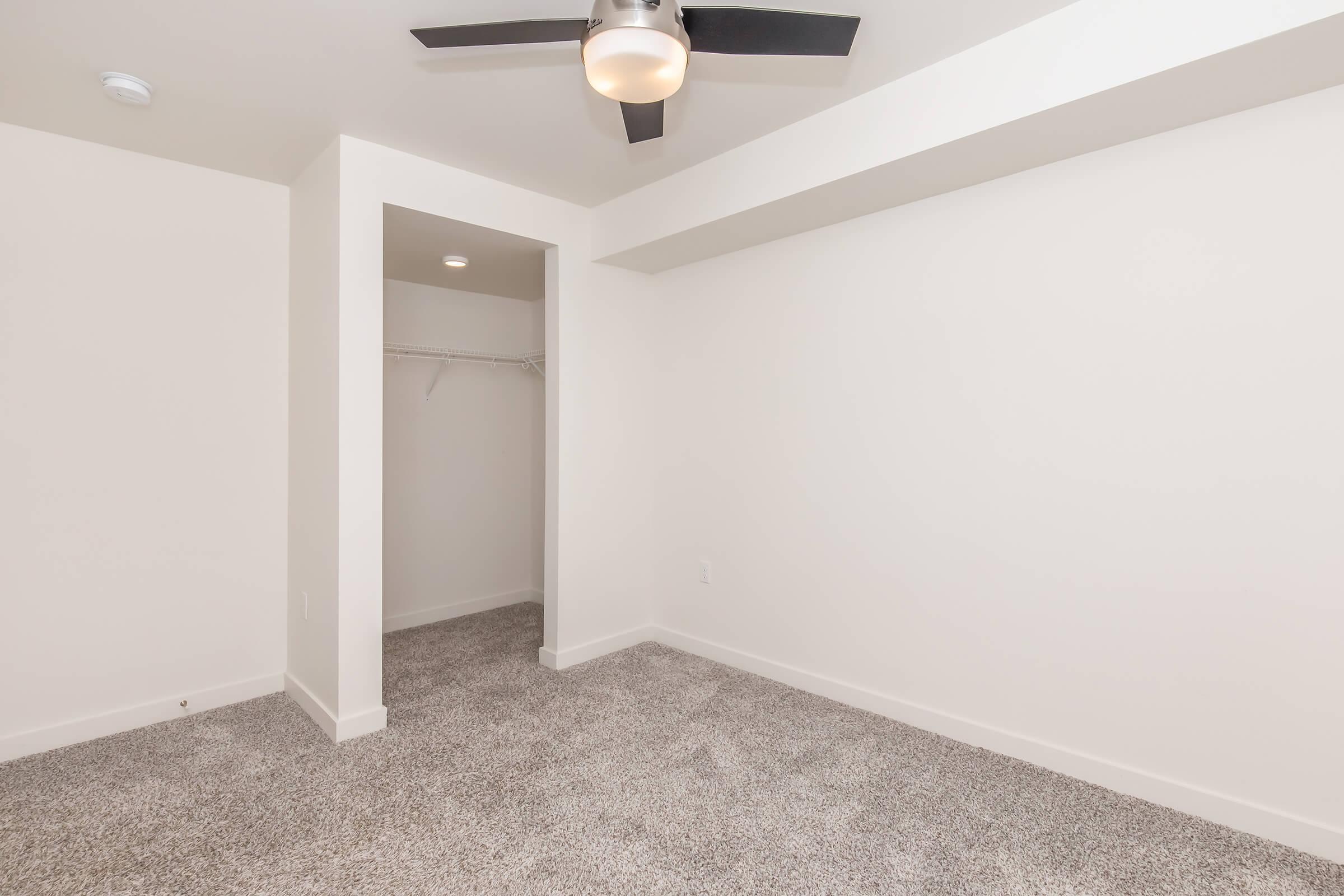 A spacious, empty room with light-colored walls and a ceiling fan. The floor is covered in carpet, and there is a small alcove or closet space on one side with a hanging rail. Natural light brightens the area, creating a clean and minimalist look.