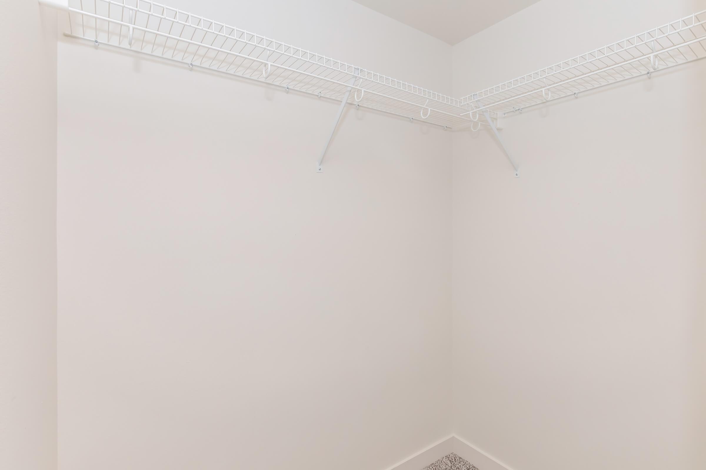 A spacious, empty closet with white walls and a wire shelving unit along the top. The floor is covered in light-colored tiles, and the space is well-lit, providing a clean and minimal appearance, ideal for storage.