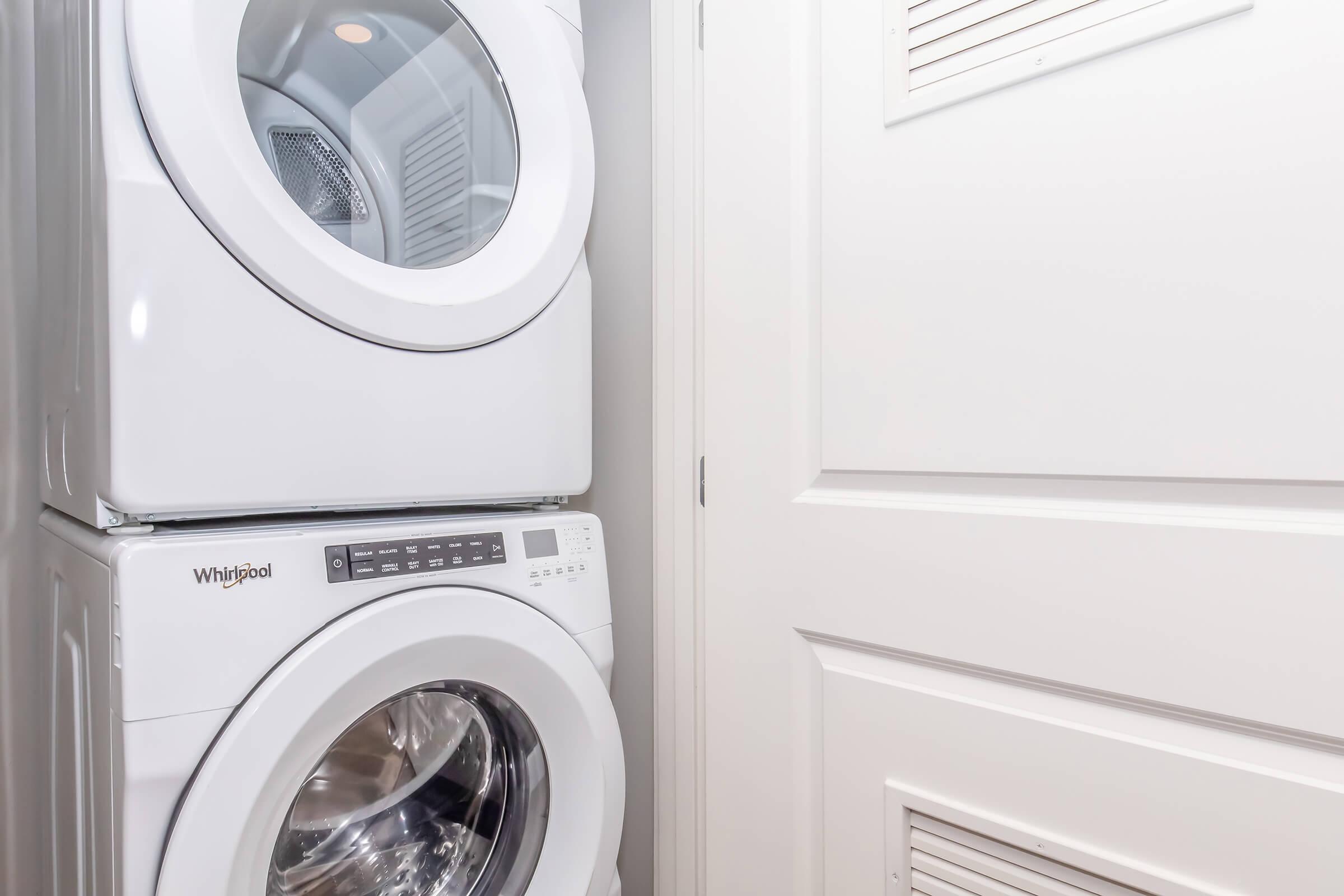 Stacked white washer and dryer appliances in a laundry room, positioned next to a closed white door. The appliances feature controls on the front panel, and the washer has a clear round door showing a stainless steel drum inside.